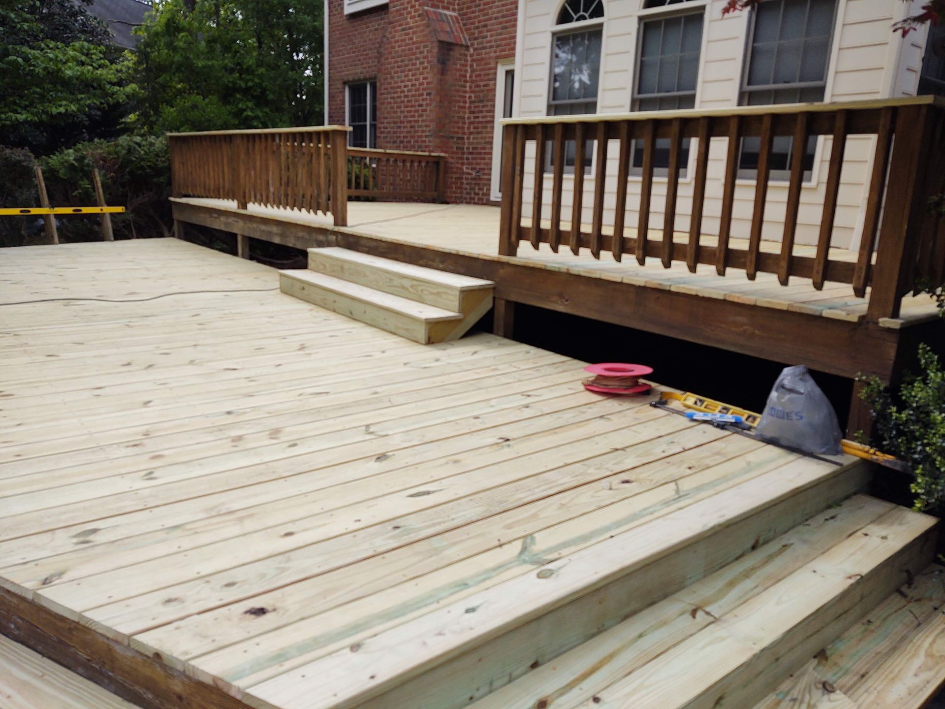 Wooden deck with steps, railings, and access to a lower area; brick house in background.