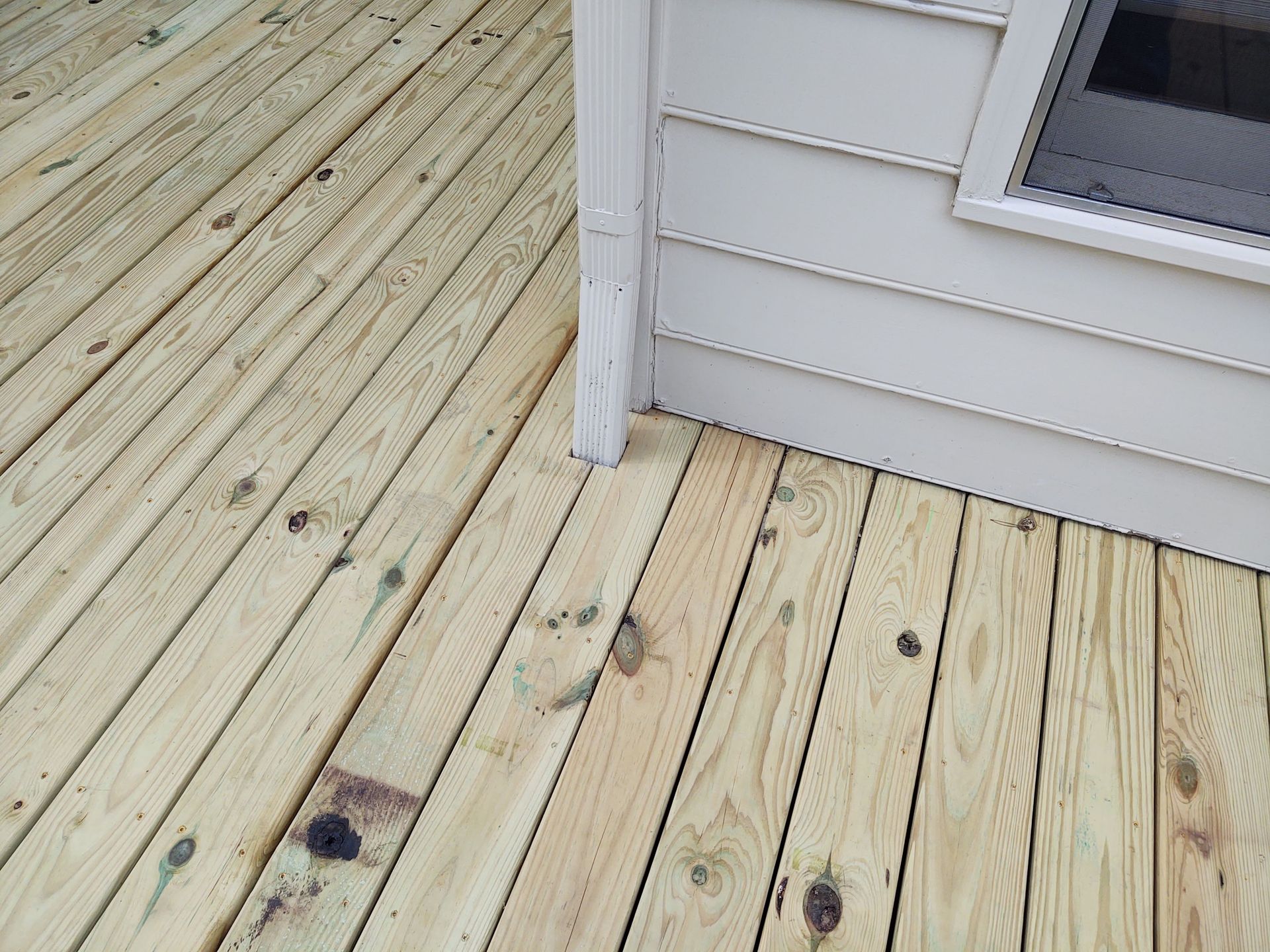Wooden deck next to a light-colored building with a window, planks in diagonal and parallel lines.