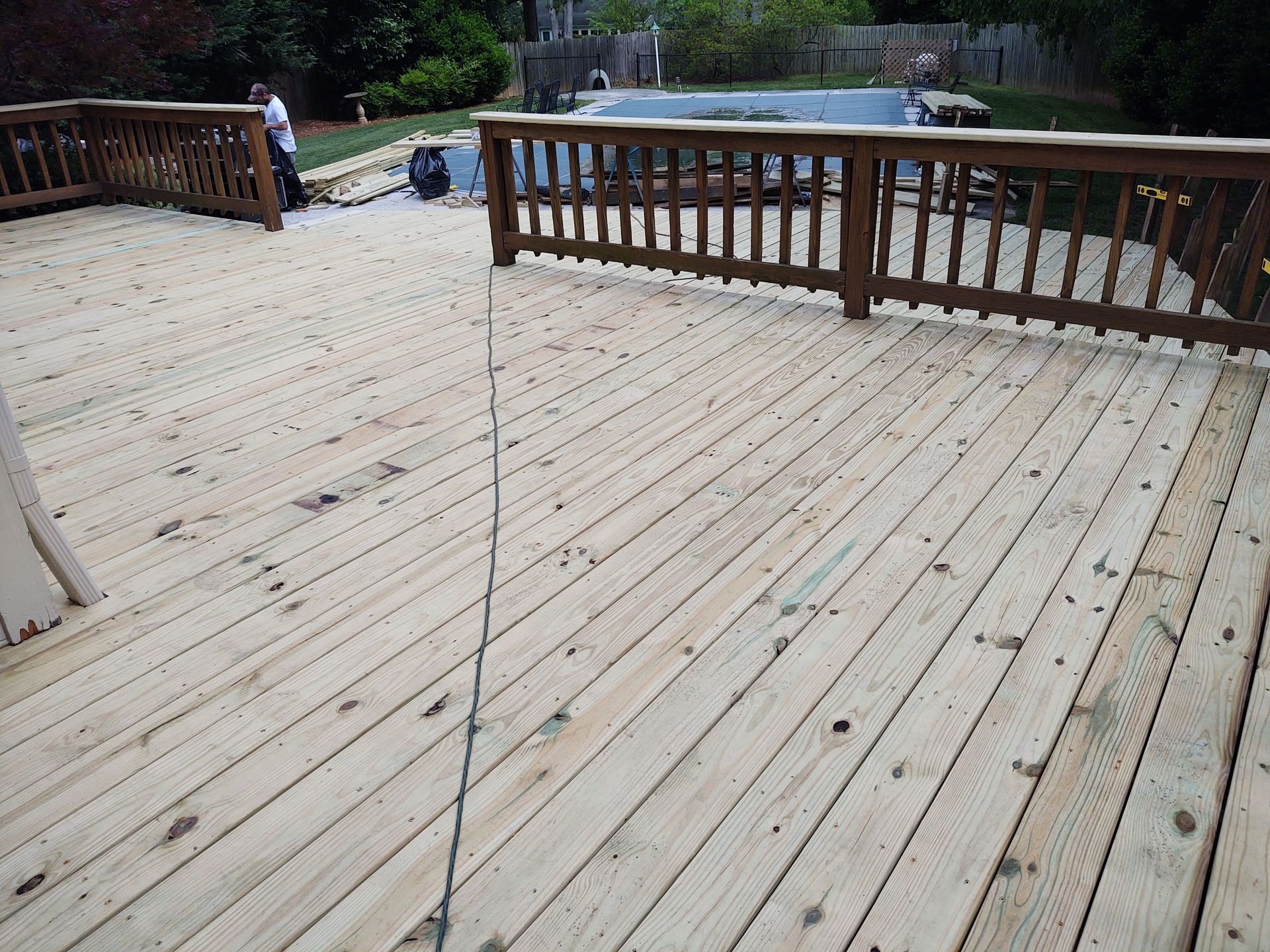 Wooden deck with railing. Person in background, pool and trees visible.