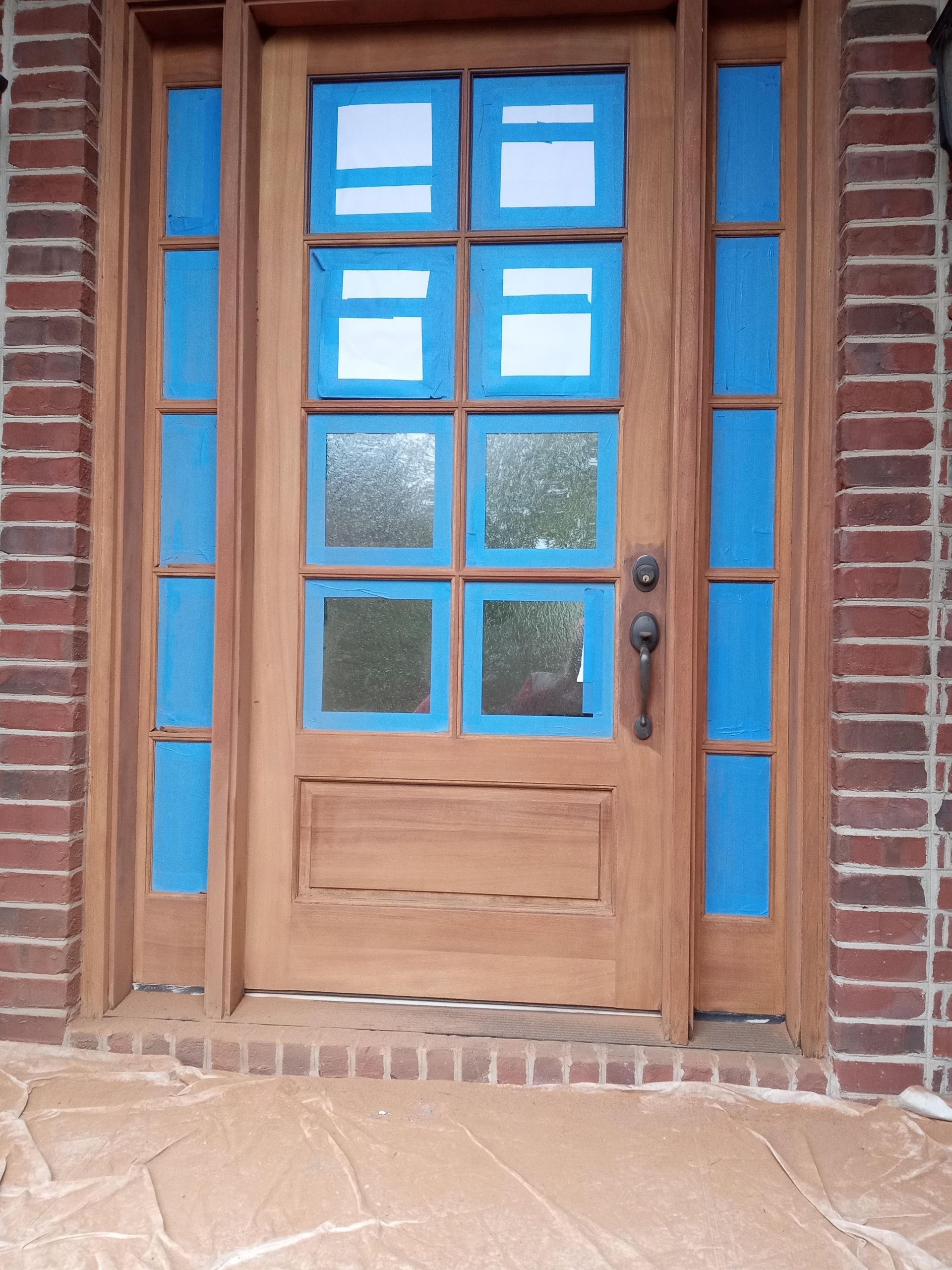 Wooden door with blue tape around the windows, ready for painting, set in a brick frame.