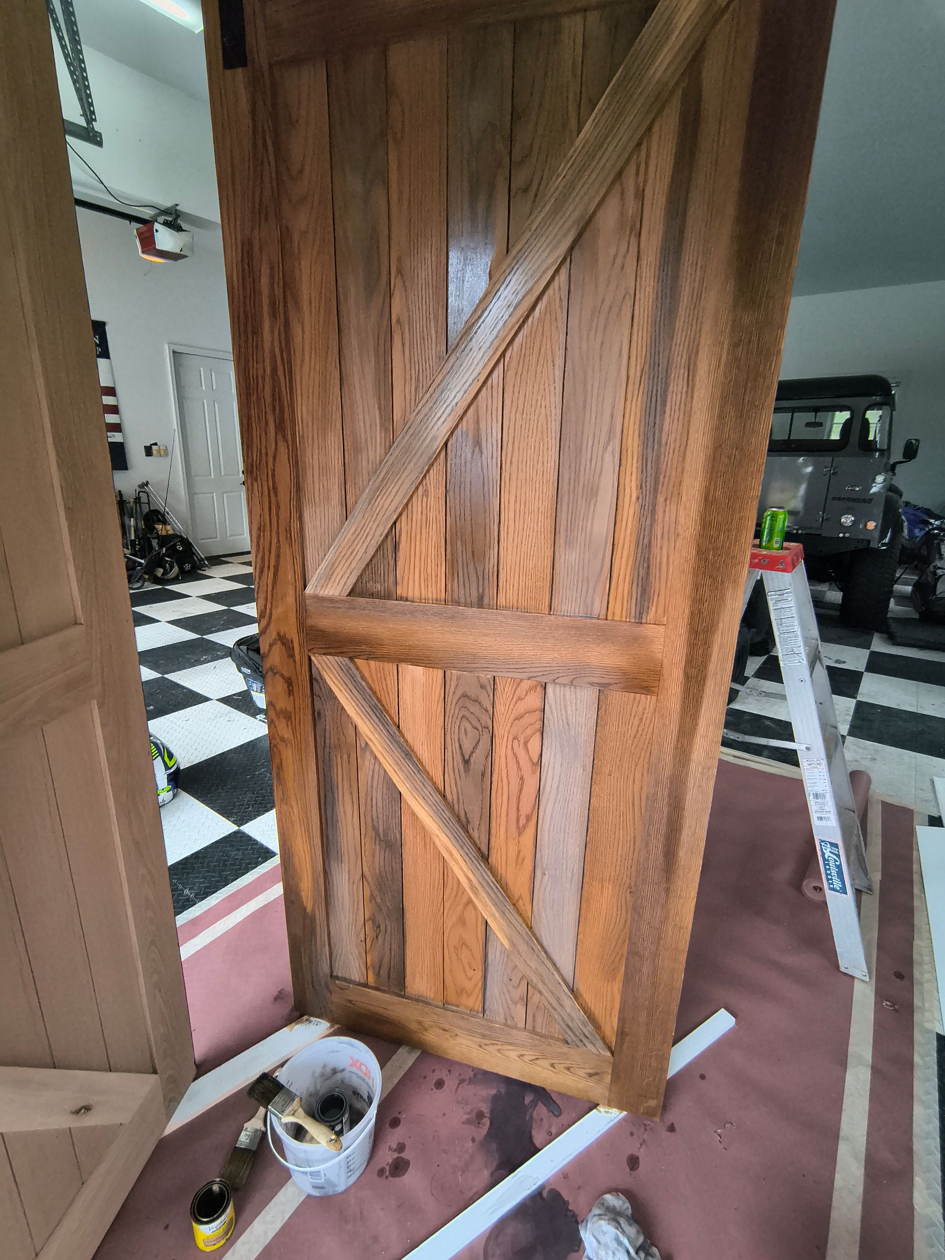 Wooden barn door with diagonal brace, stained in shades of brown, resting on a rug in a garage.