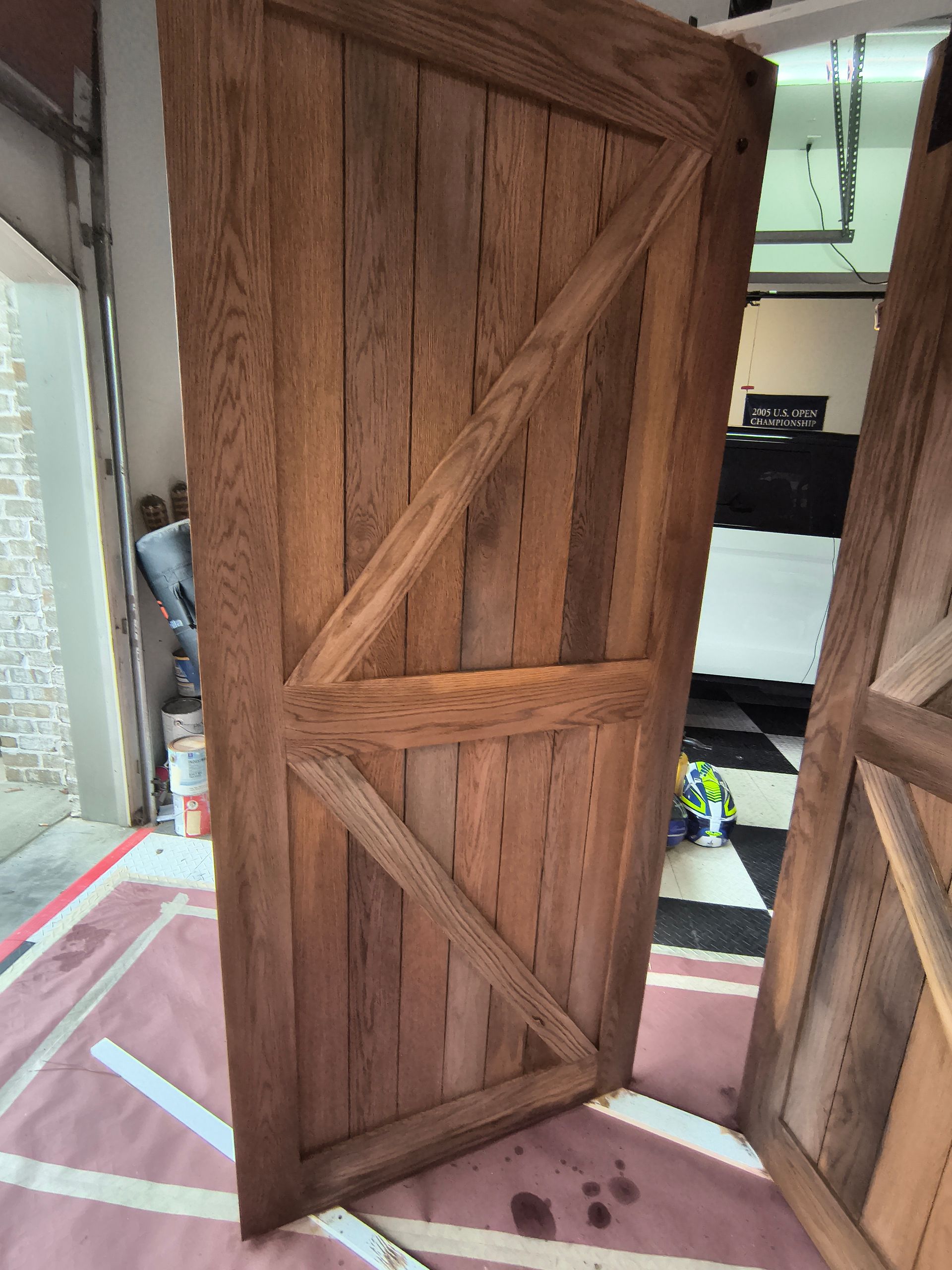 Two stained wooden doors leaning on a red mat, against a background of a garage and a checkered floor.