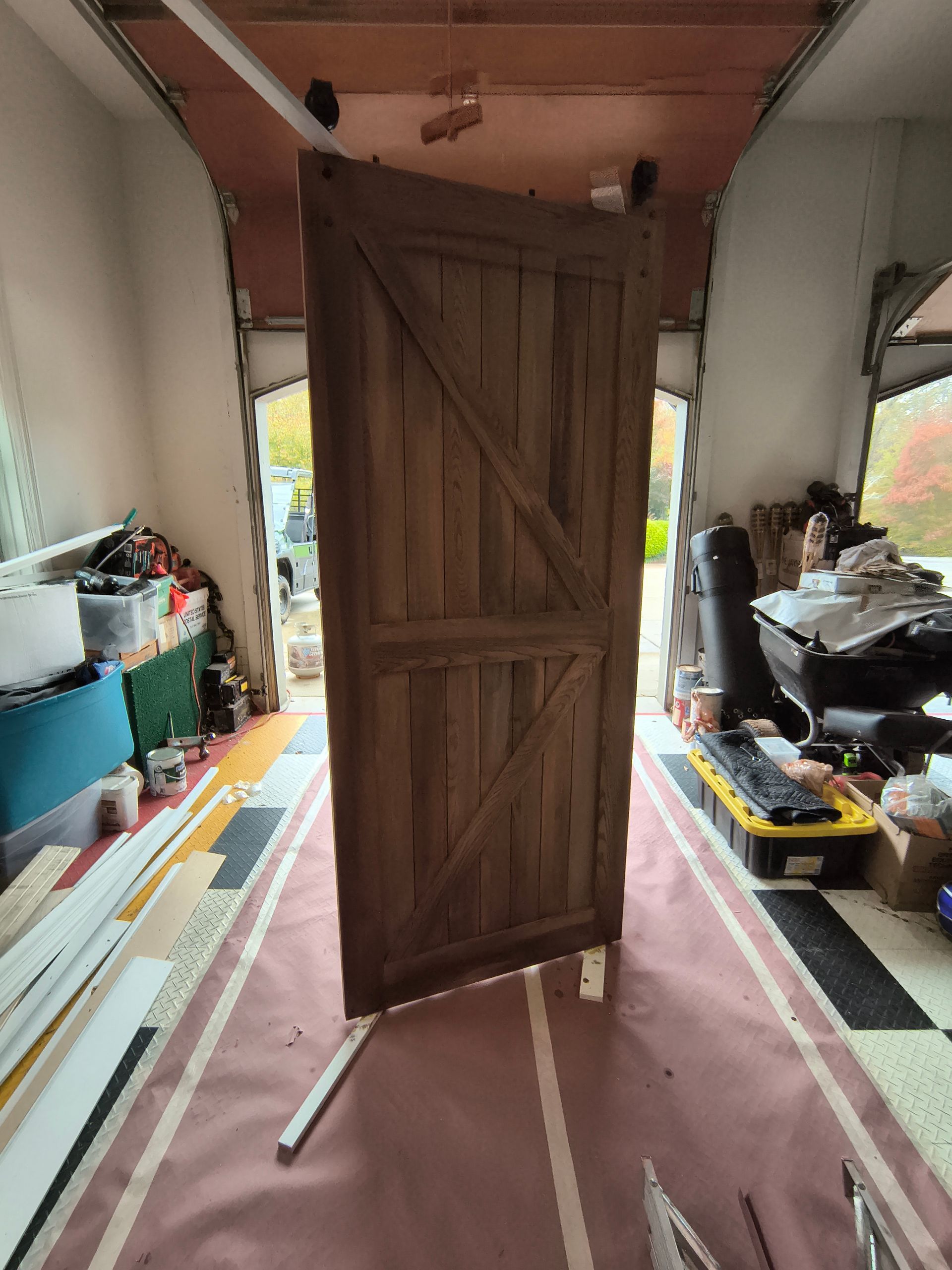 Wooden door inside a garage, resting on supports. Diagonal brace, brown planks, overhead track.