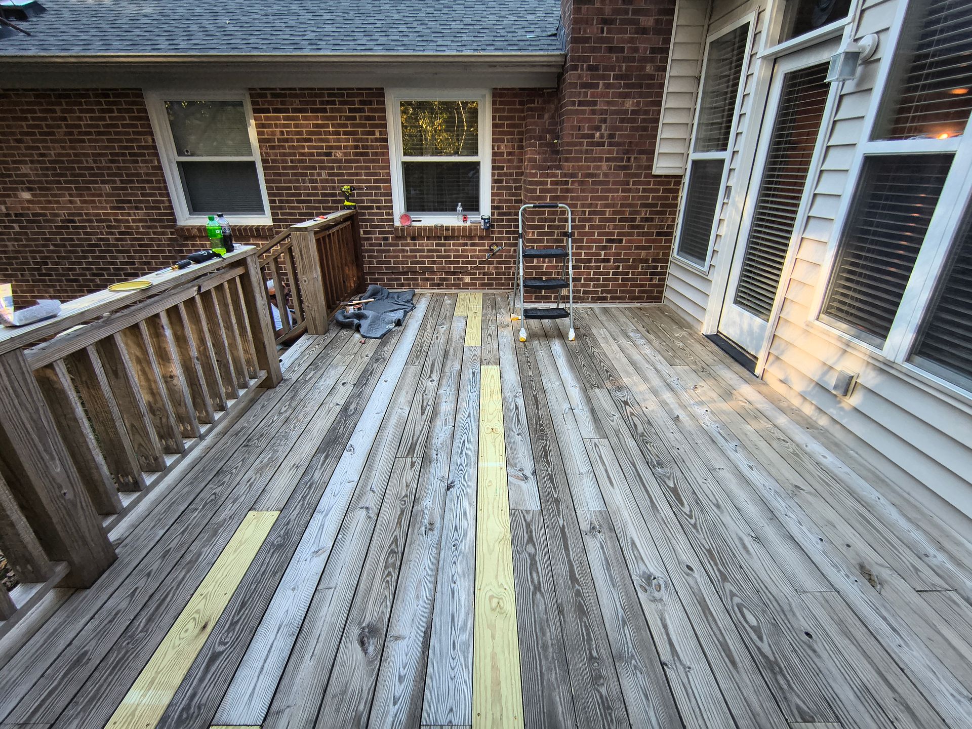 Deck with some planks removed and replaced with new wood, next to a brick house.
