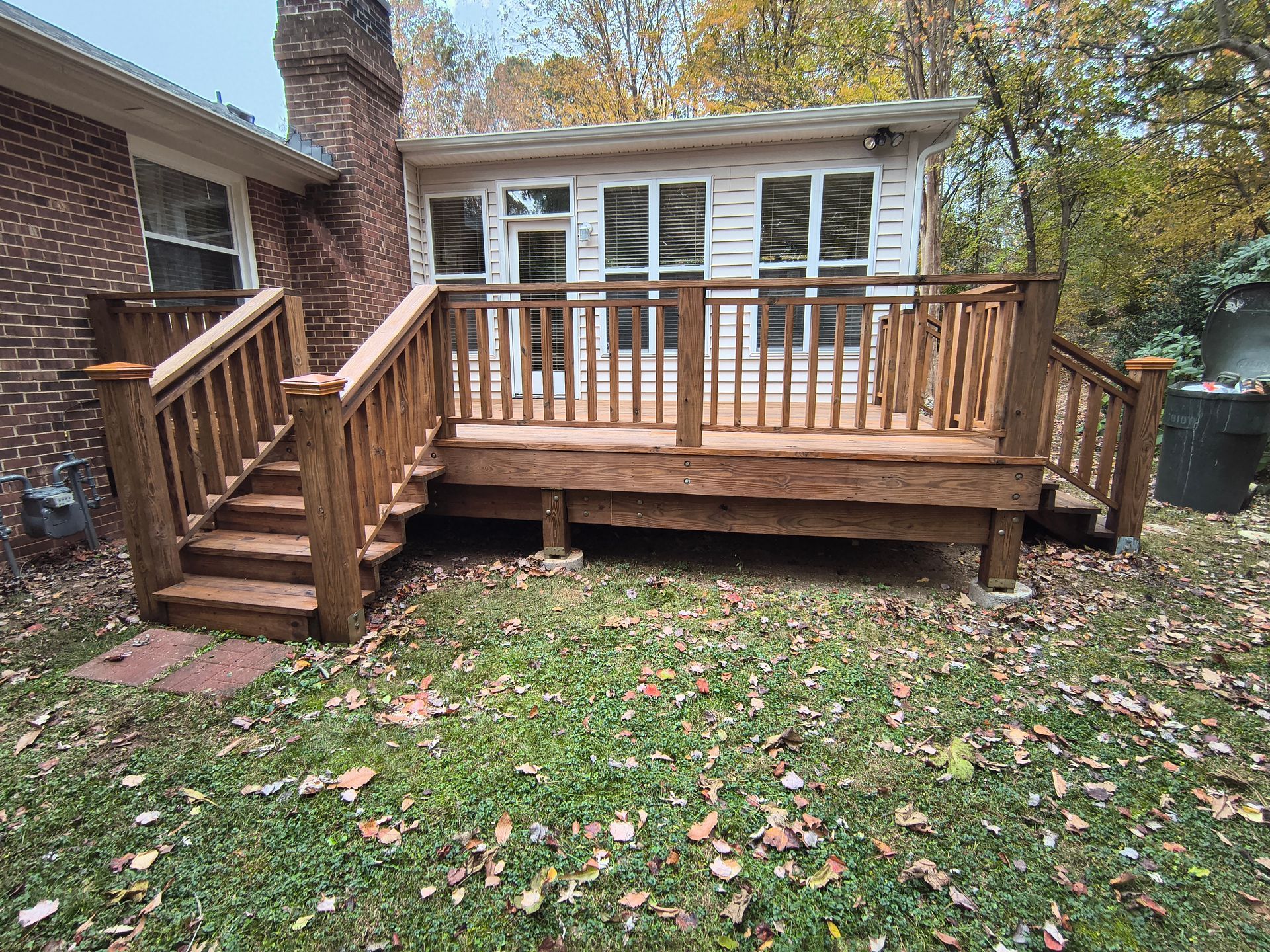 Wooden deck with railing, stairs, and attached to a brick house with a sunroom in the background.