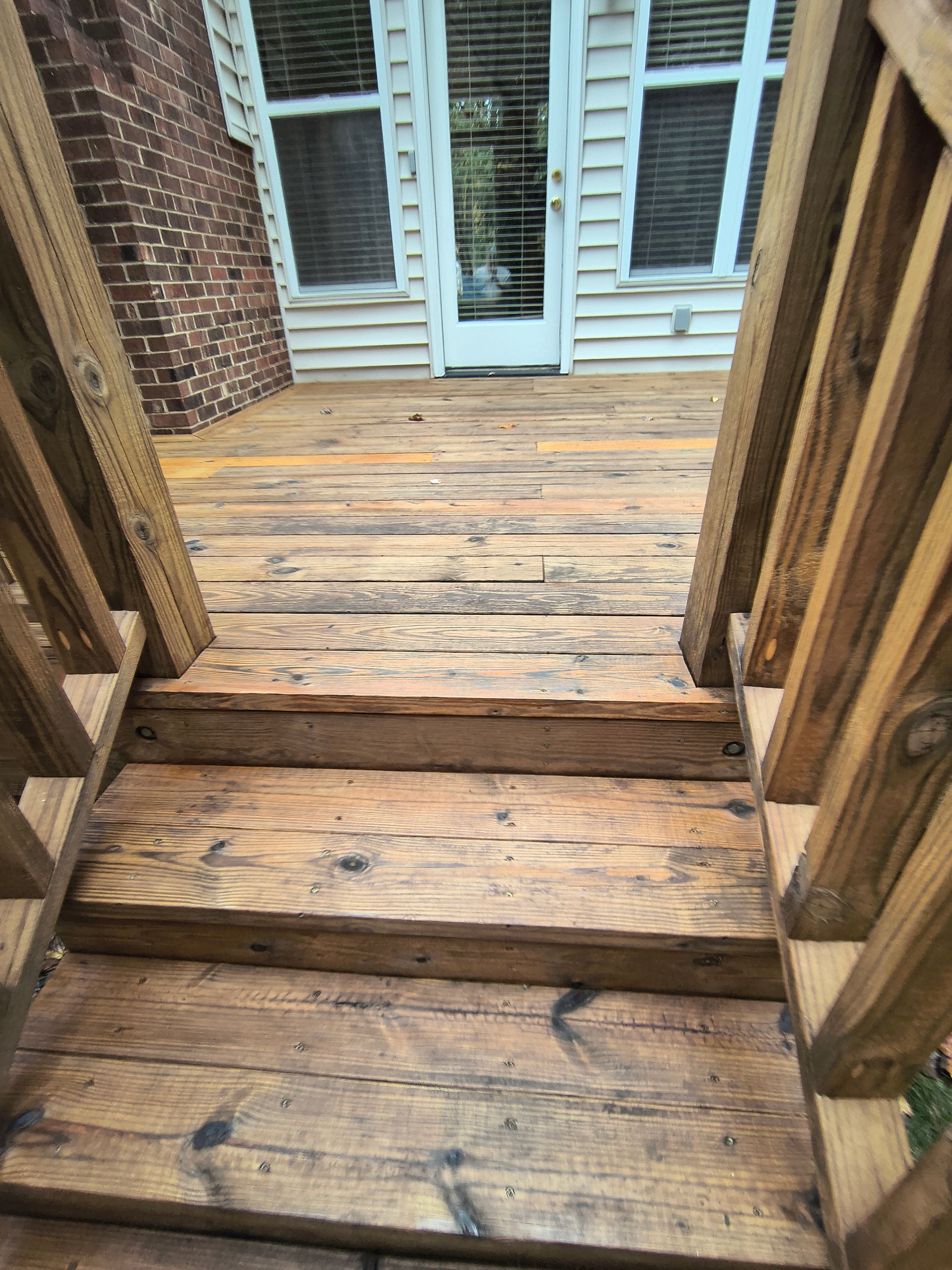 Wooden deck with stairs leading to a back door. Faded wood, brick building in background.