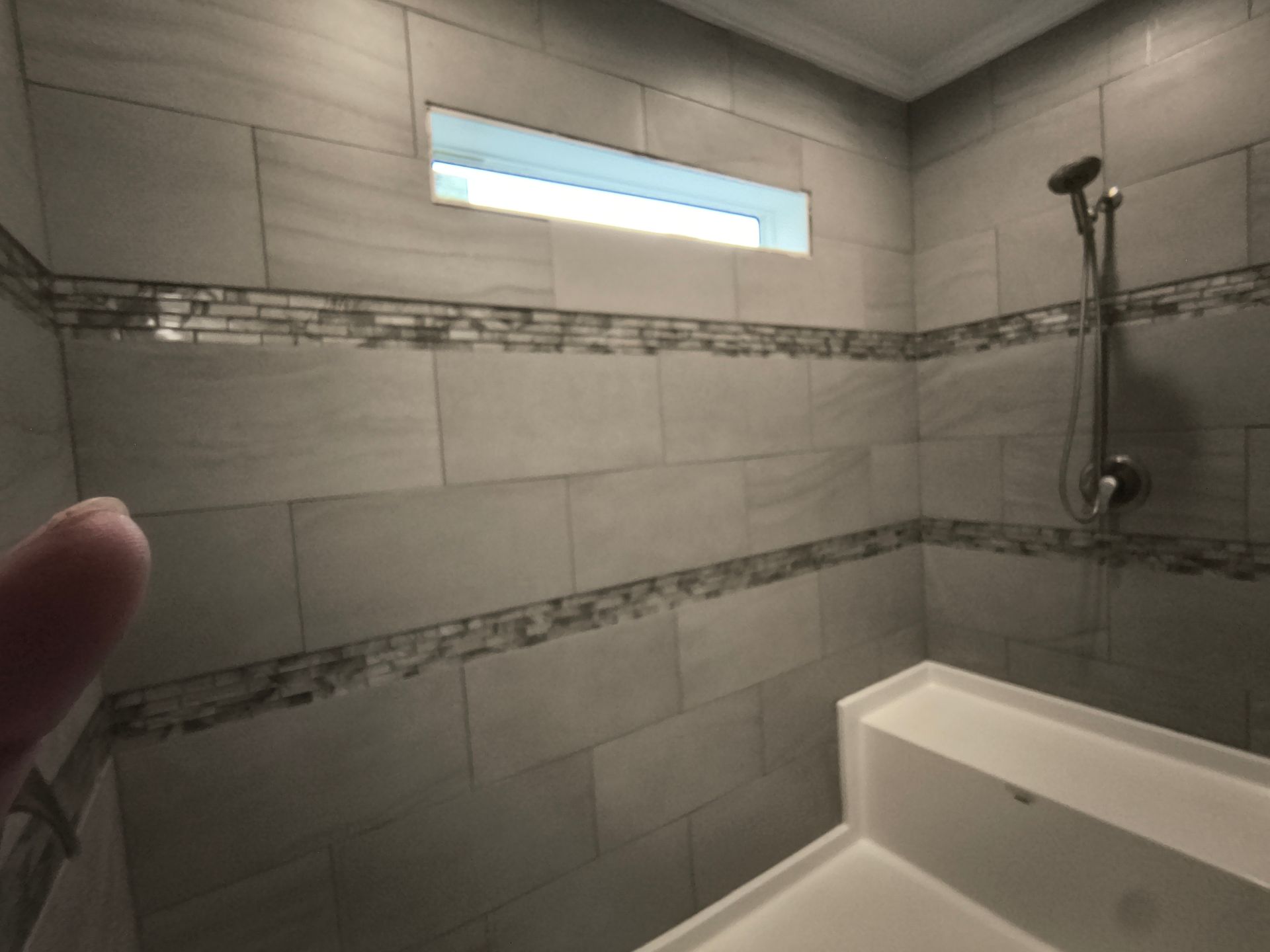 Bathroom interior with gray tile walls, mosaic tile border, showerhead, built-in bench, and a small window.