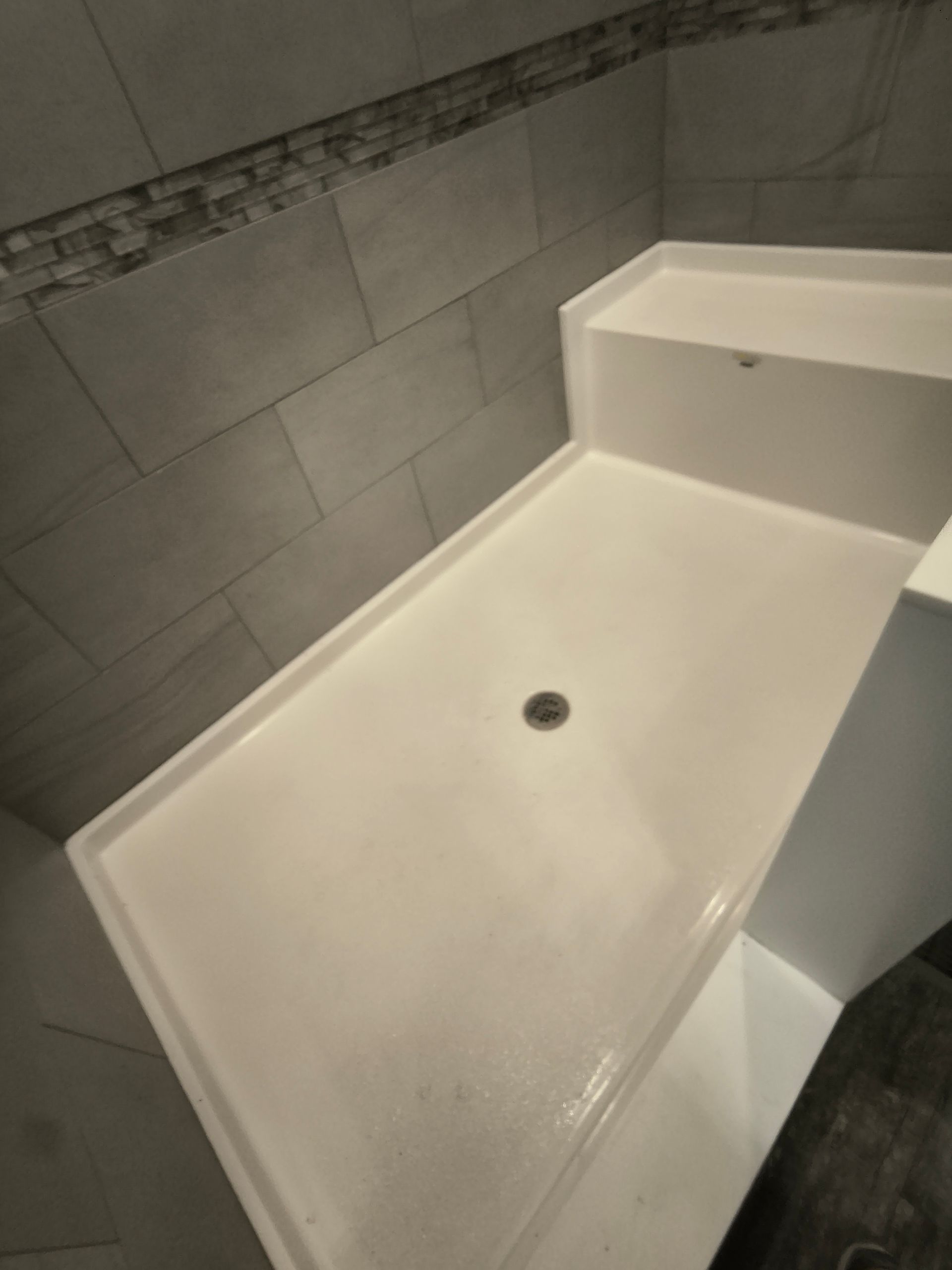 White shower with a built-in bench, gray tile walls, and a drain.