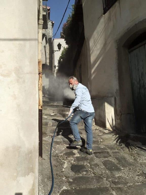 Un uomo con la mascherina spruzza acqua con un tubo in uno stretto vicolo acciottolato, creando una nebbia.