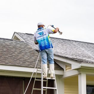Person using a high-pressure washer while standing on a ladder to clean the metal roof of a house.