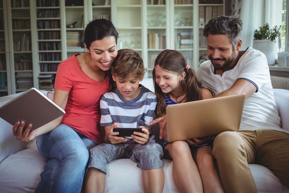 A family is sitting on a couch using a laptop and a tablet.