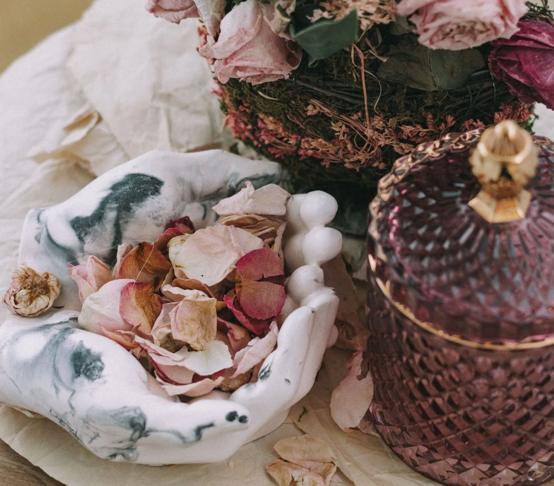 A deep purple textured glass canister sits on a table surrounded by a hands-shaped bowl filled with a fragrant potpourri of flower petals, and a dried floral bouquet.