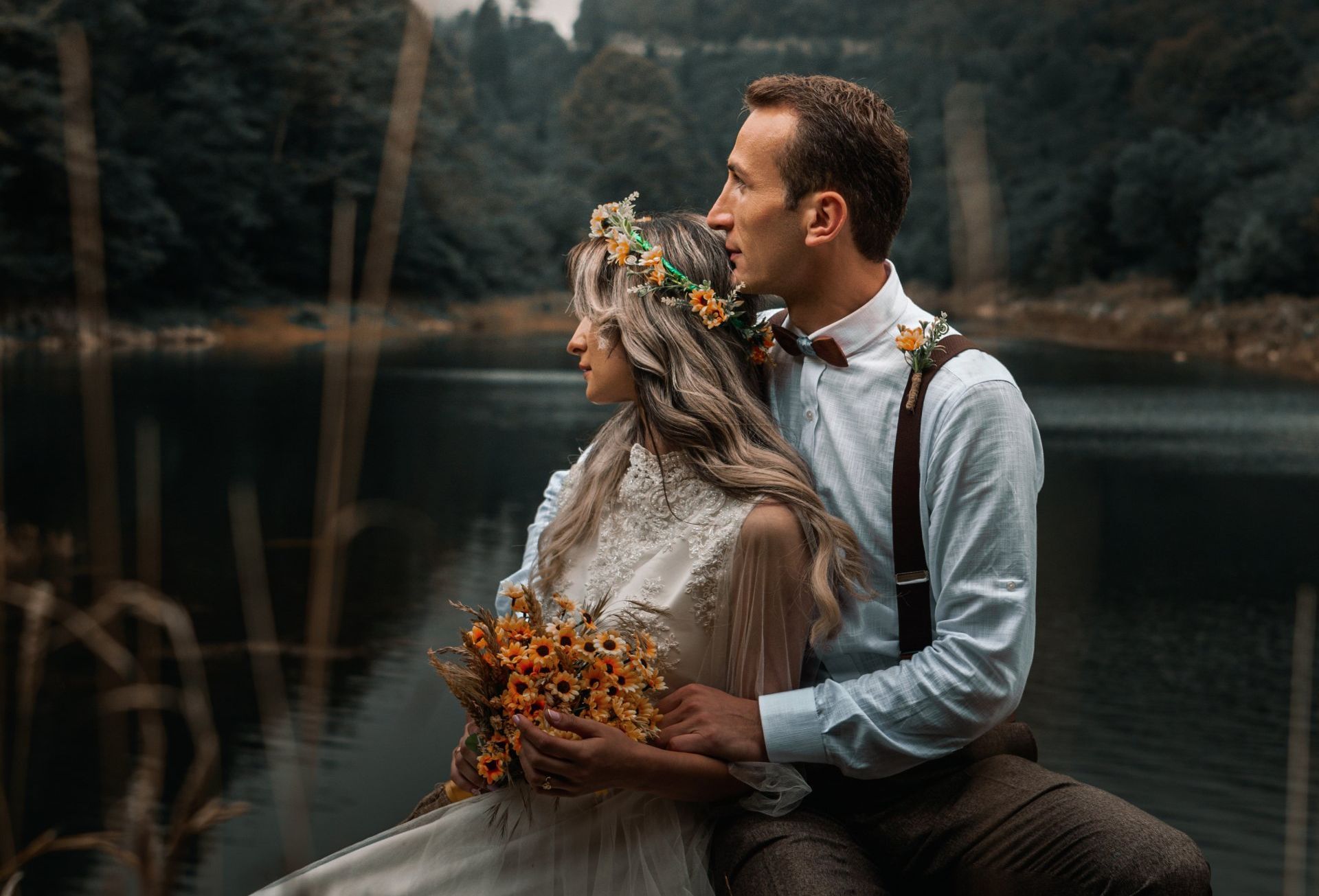 A couple sits by a lake, where the bride, adorned with an orange flower crown, is held by her groom.