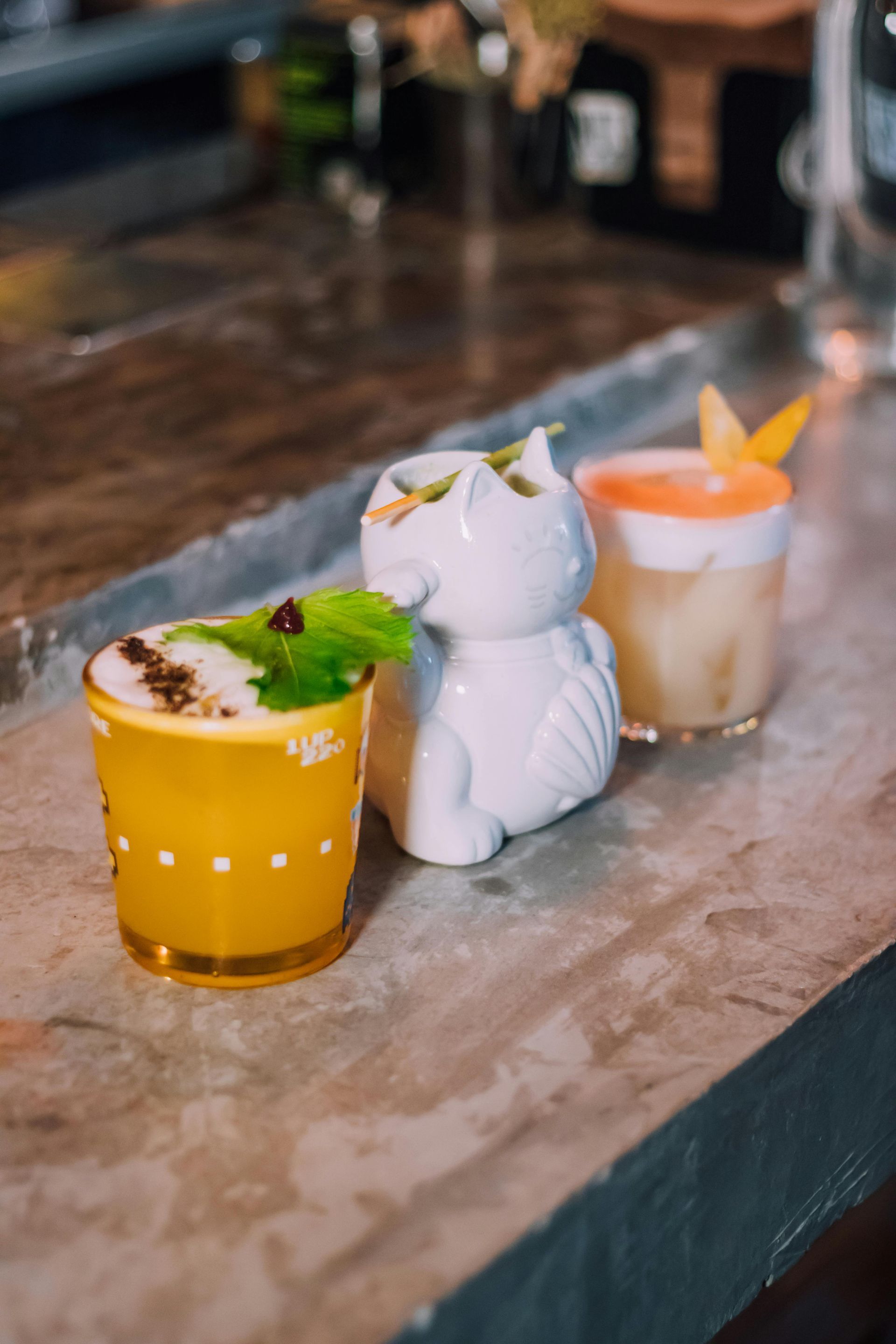 Three cocktails on a bar, one in an owl-shaped glass, yellow and cream colors, with garnish.