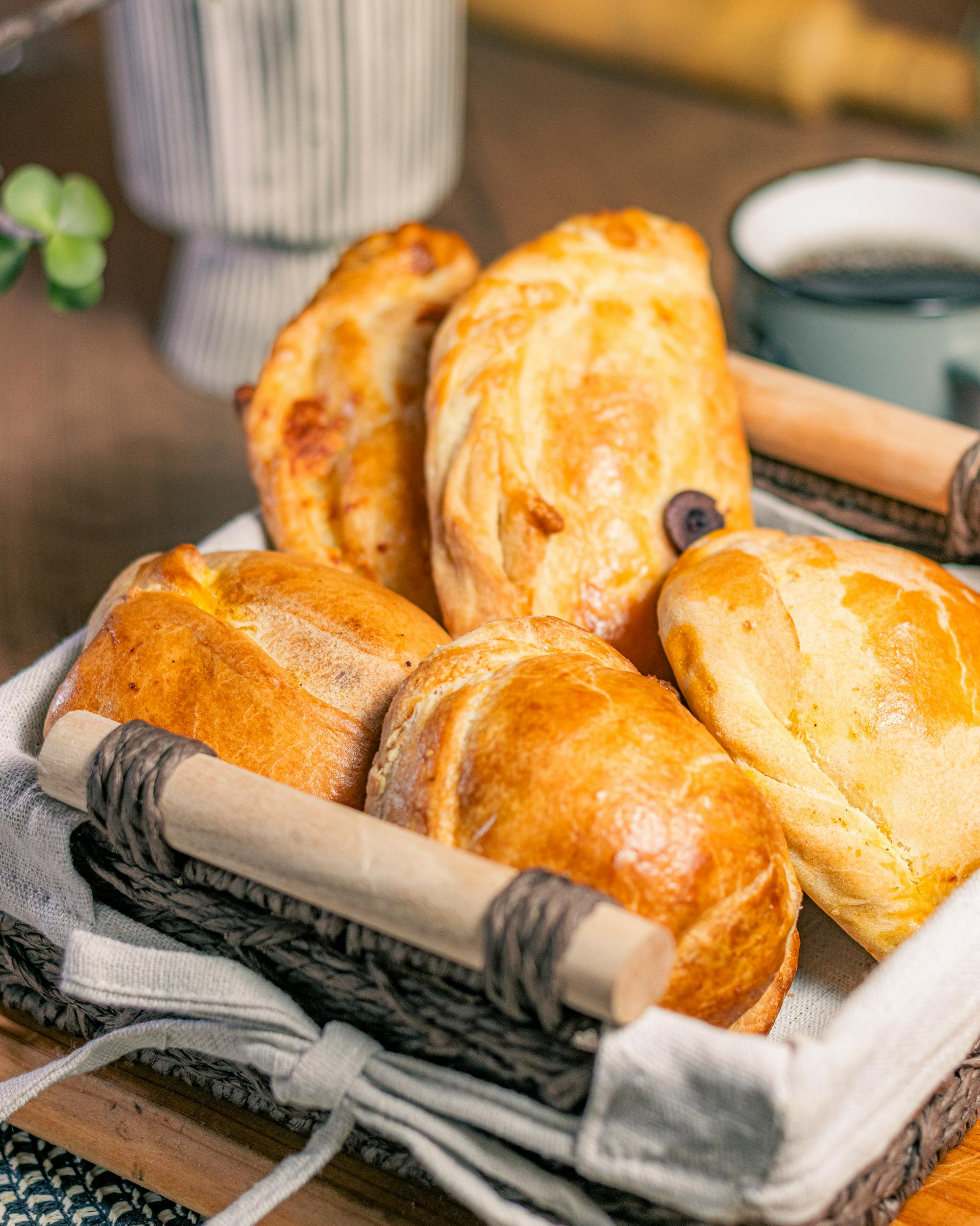 Basket of golden-brown baked pastries, resting on a white cloth, with a cup and rolling pin nearby.