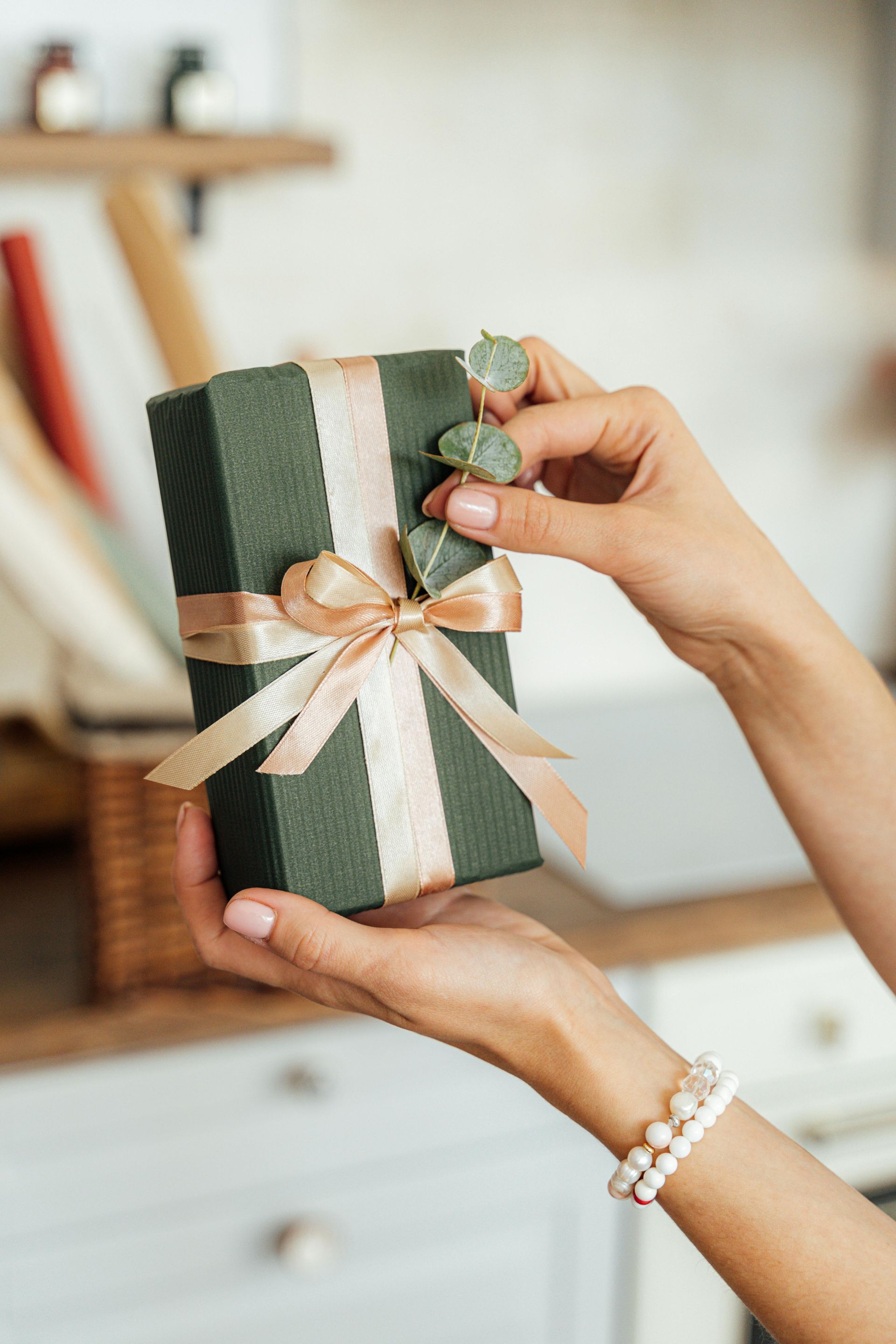 Hands holding a wrapped green gift box with a beige ribbon and small green decorations.