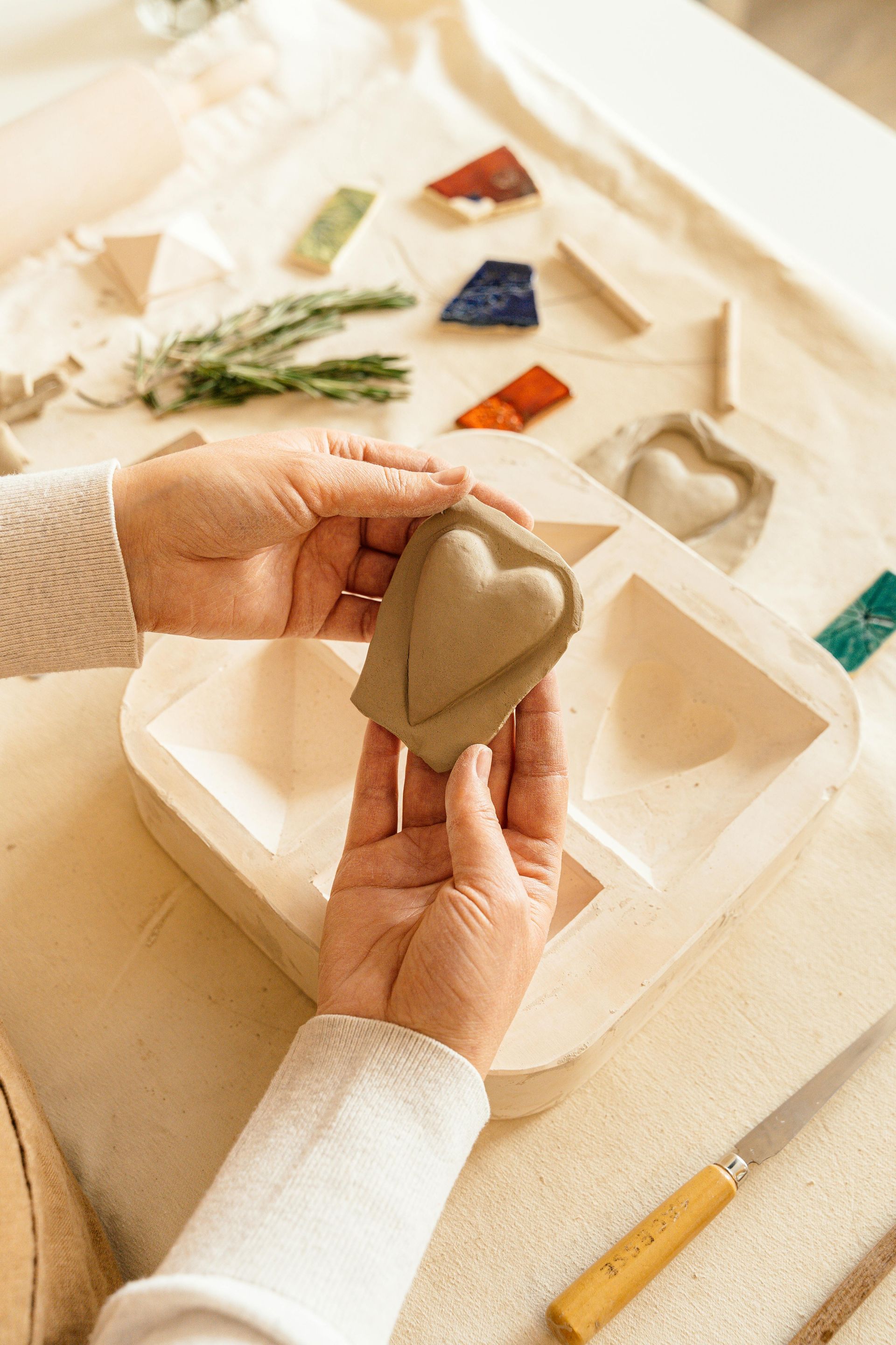 Hands holding clay heart mold over a tray, with craft supplies on a table.