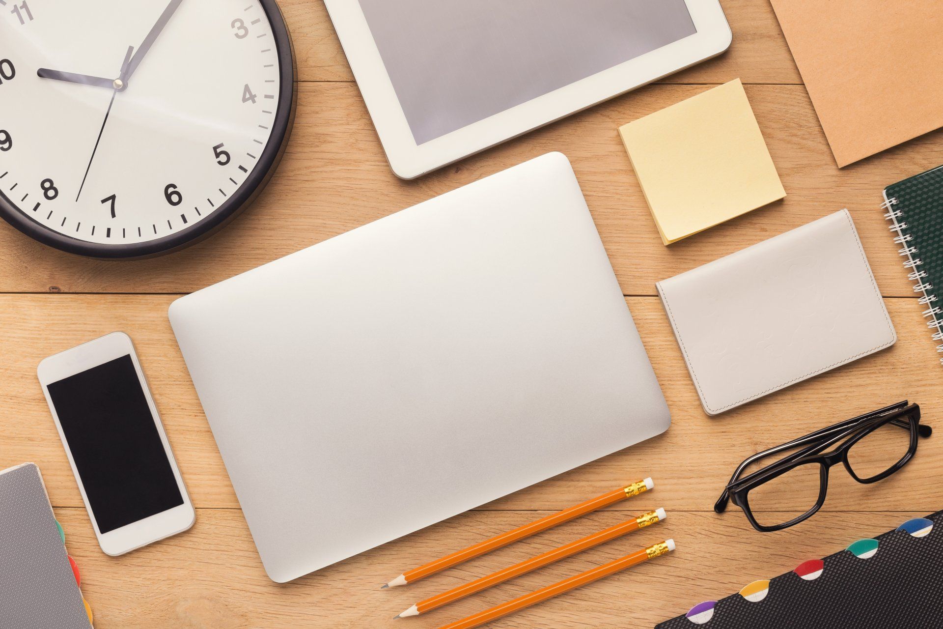 A wooden desk with a laptop , tablet , cell phone , pencils , glasses and a clock.