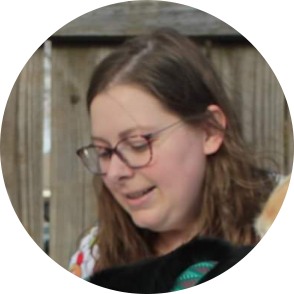 Woman with glasses looking down, light skin, brown hair, seated outdoors.