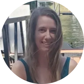 Woman with long brown hair smiles outdoors in front of a body of water.