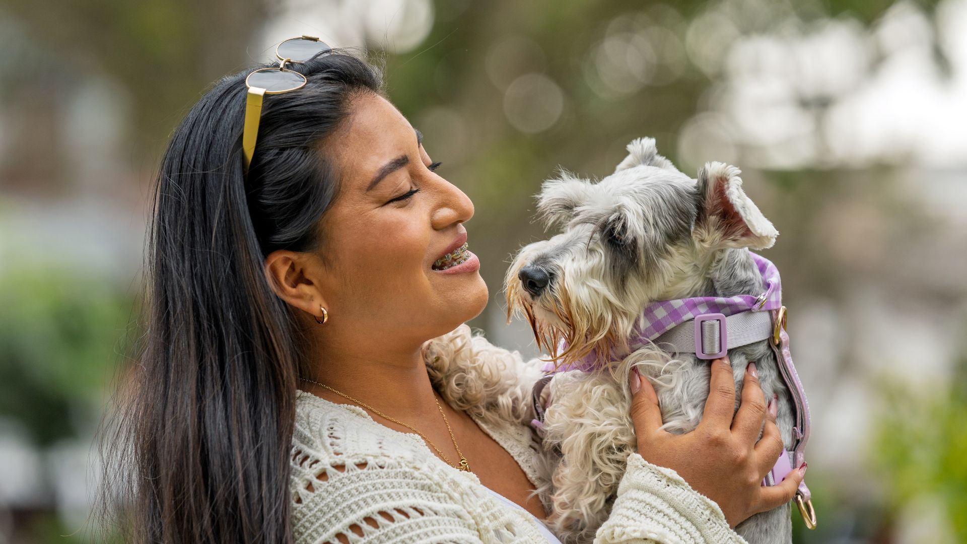 Woman holding a small, gray dog; both are smiling outside.