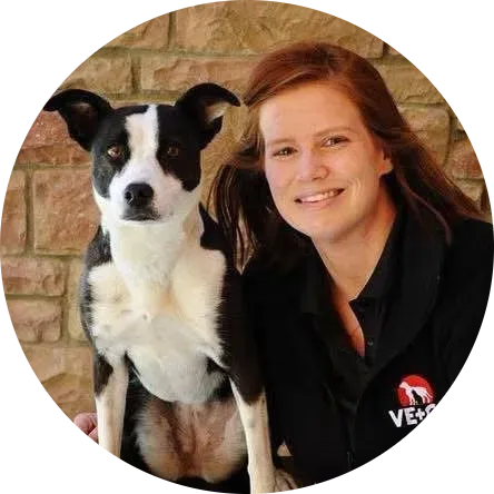 Woman in black coat smiles next to a black and white dog outdoors, brick wall background.