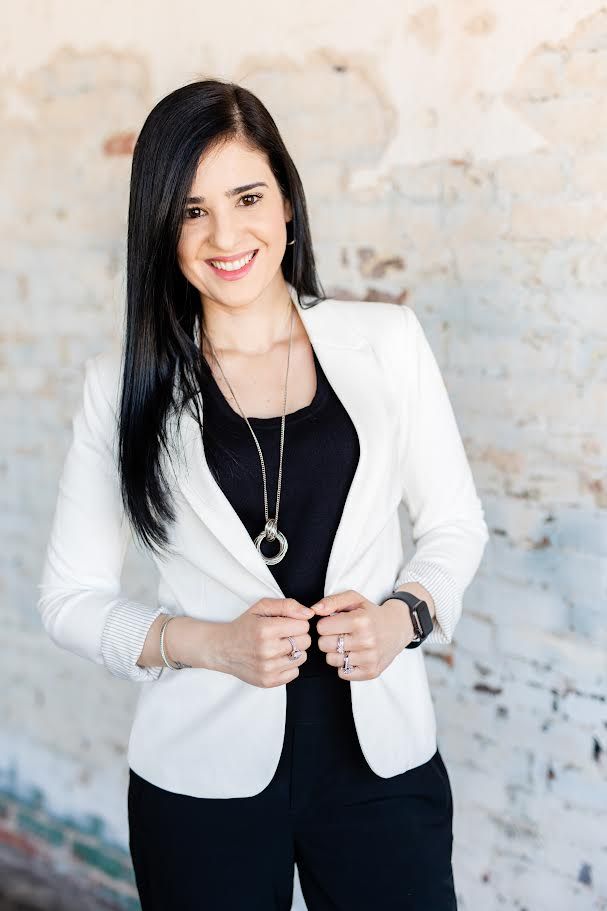 Woman smiling, wearing a white blazer and black shirt, outdoors, soft lighting.