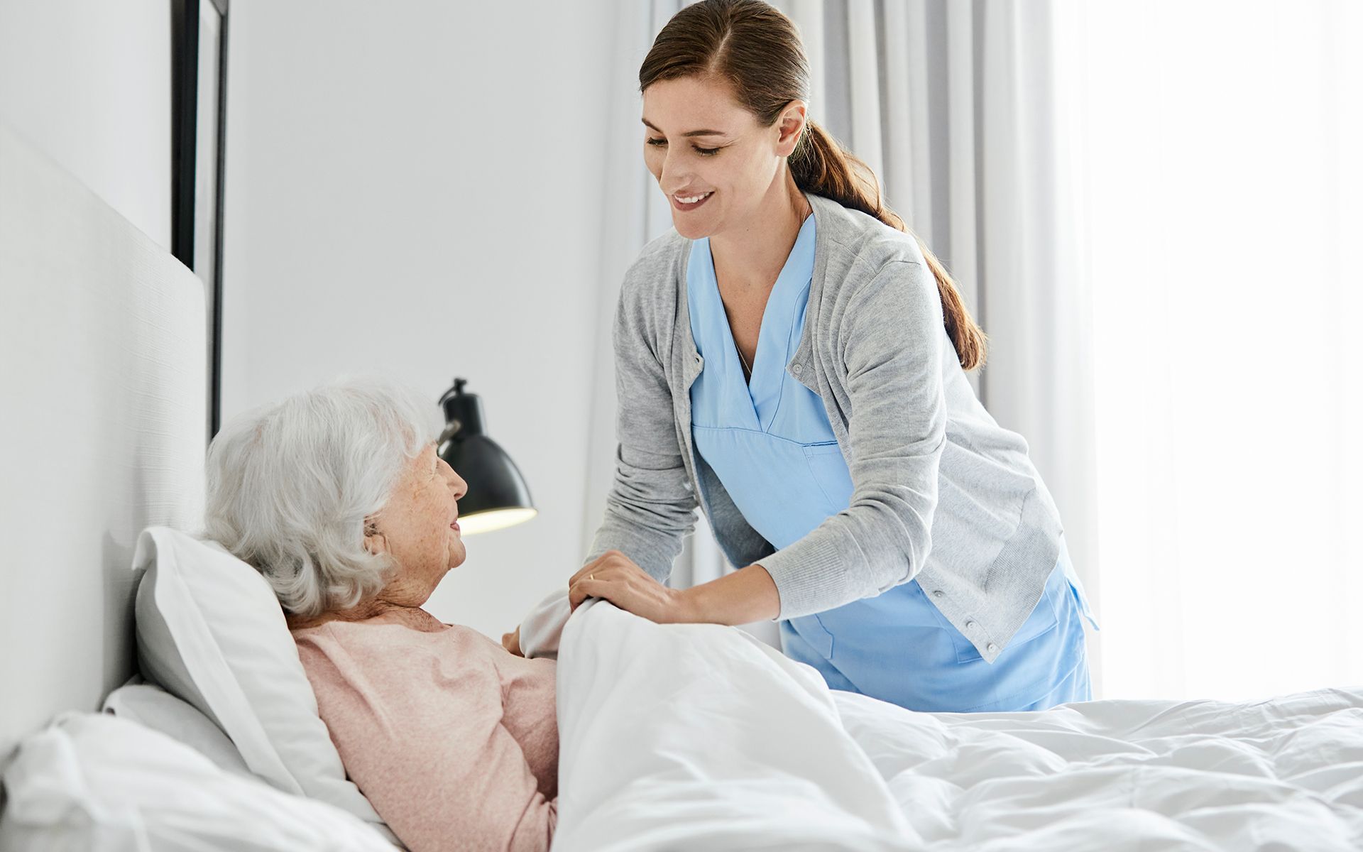 A nurse is talking to an elderly woman laying in a bed.