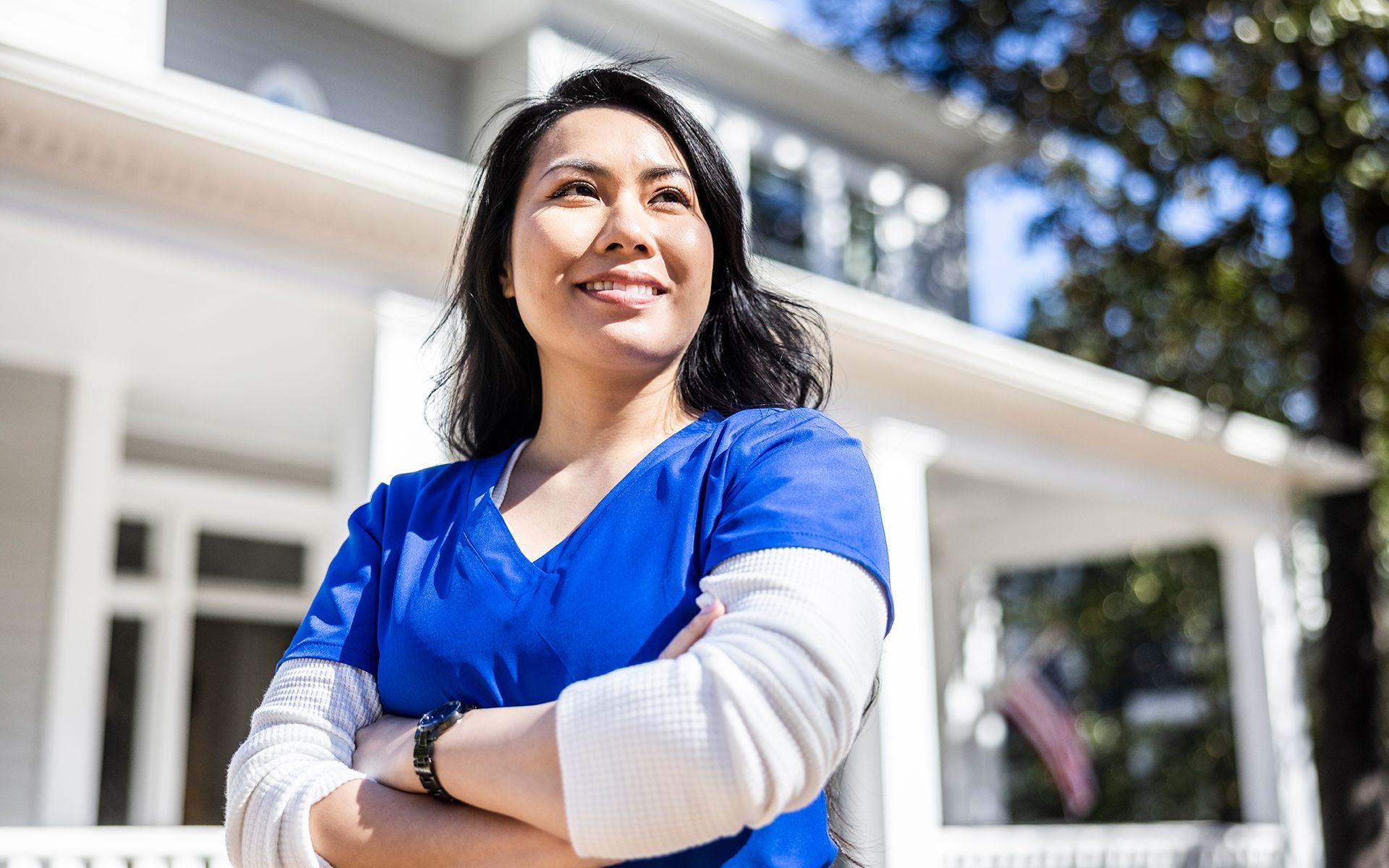 A woman in a blue scrub is standing in front of a house with her arms crossed.