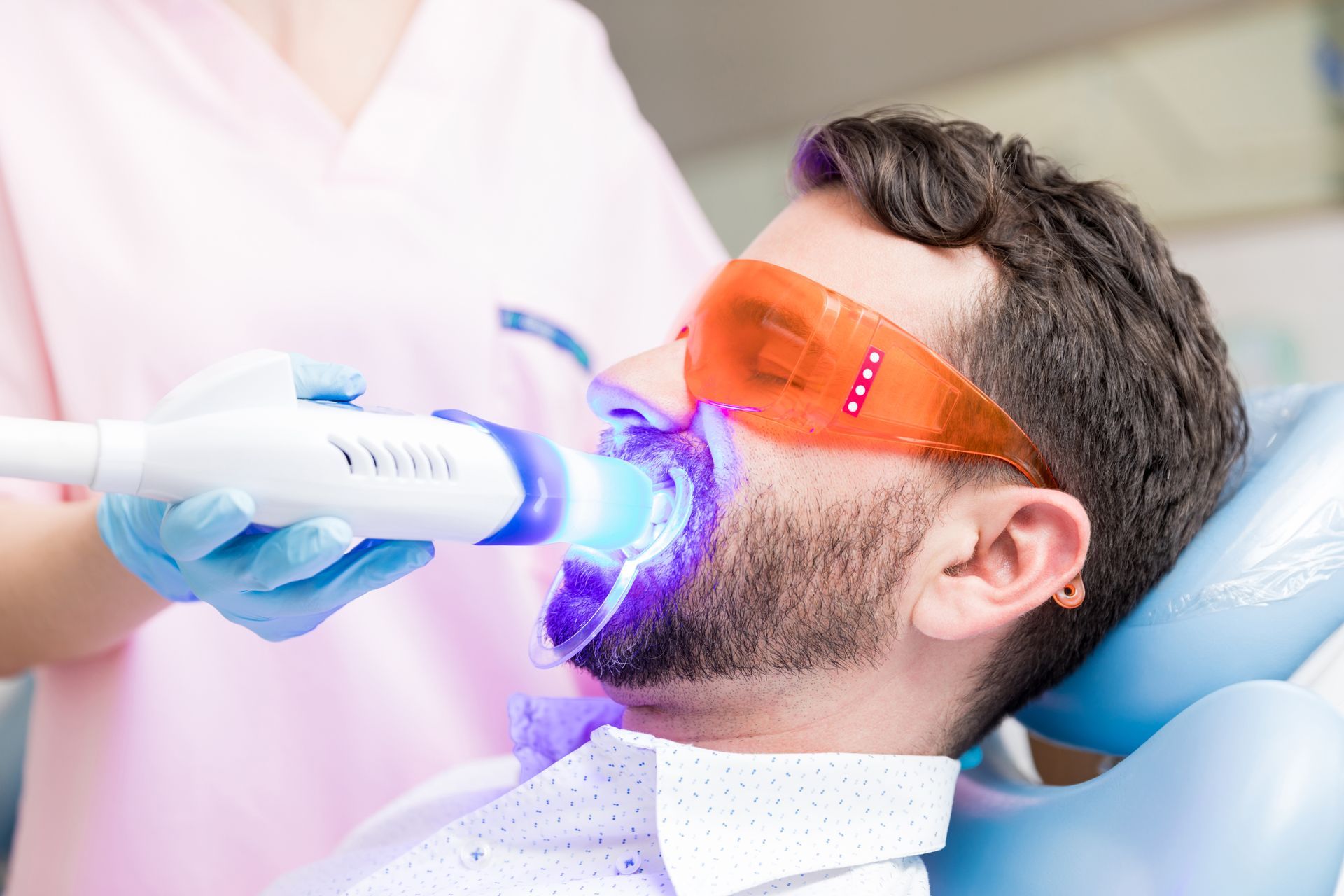 Man undergoing teeth whitening with blue light in a dental office.