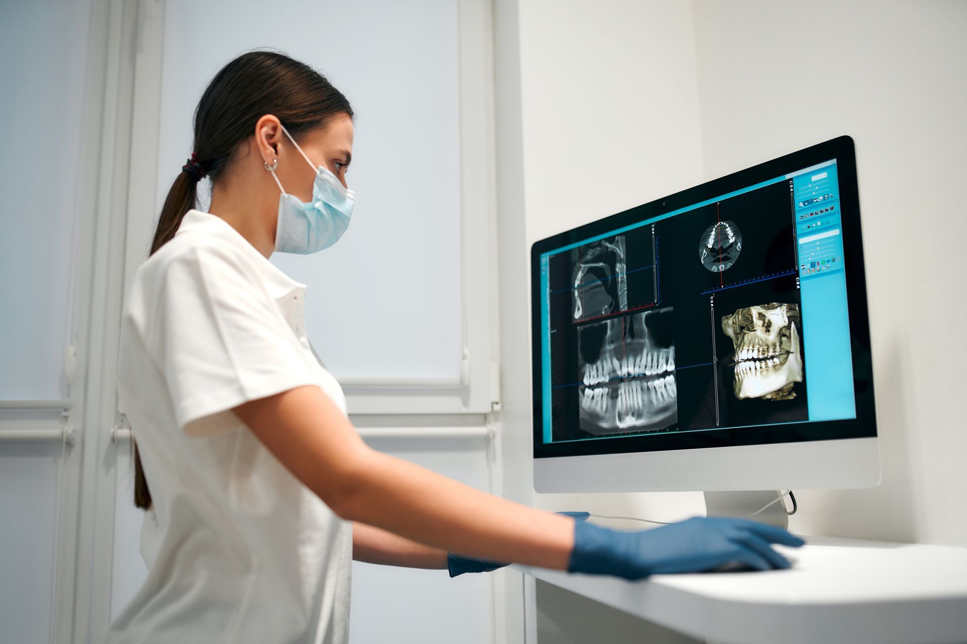 Dentist in mask and gloves reviews a dental X-ray on a computer screen.