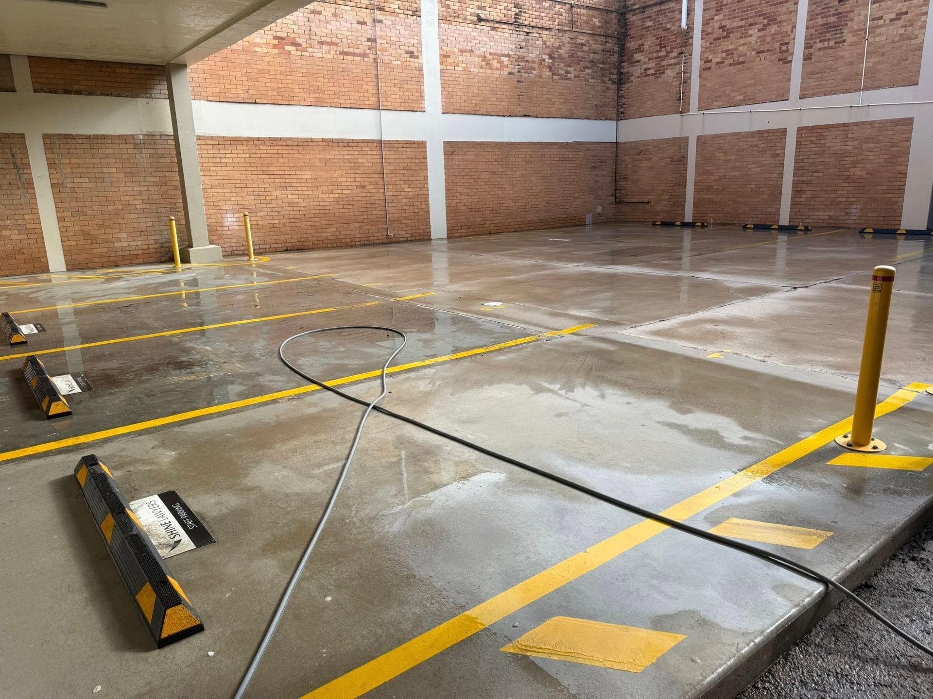 A wet, paved parking garage with yellow painted lines, parking stops, and a brick wall in the background. — CPR Exterior Cleaning in Bundaberg West, QLD