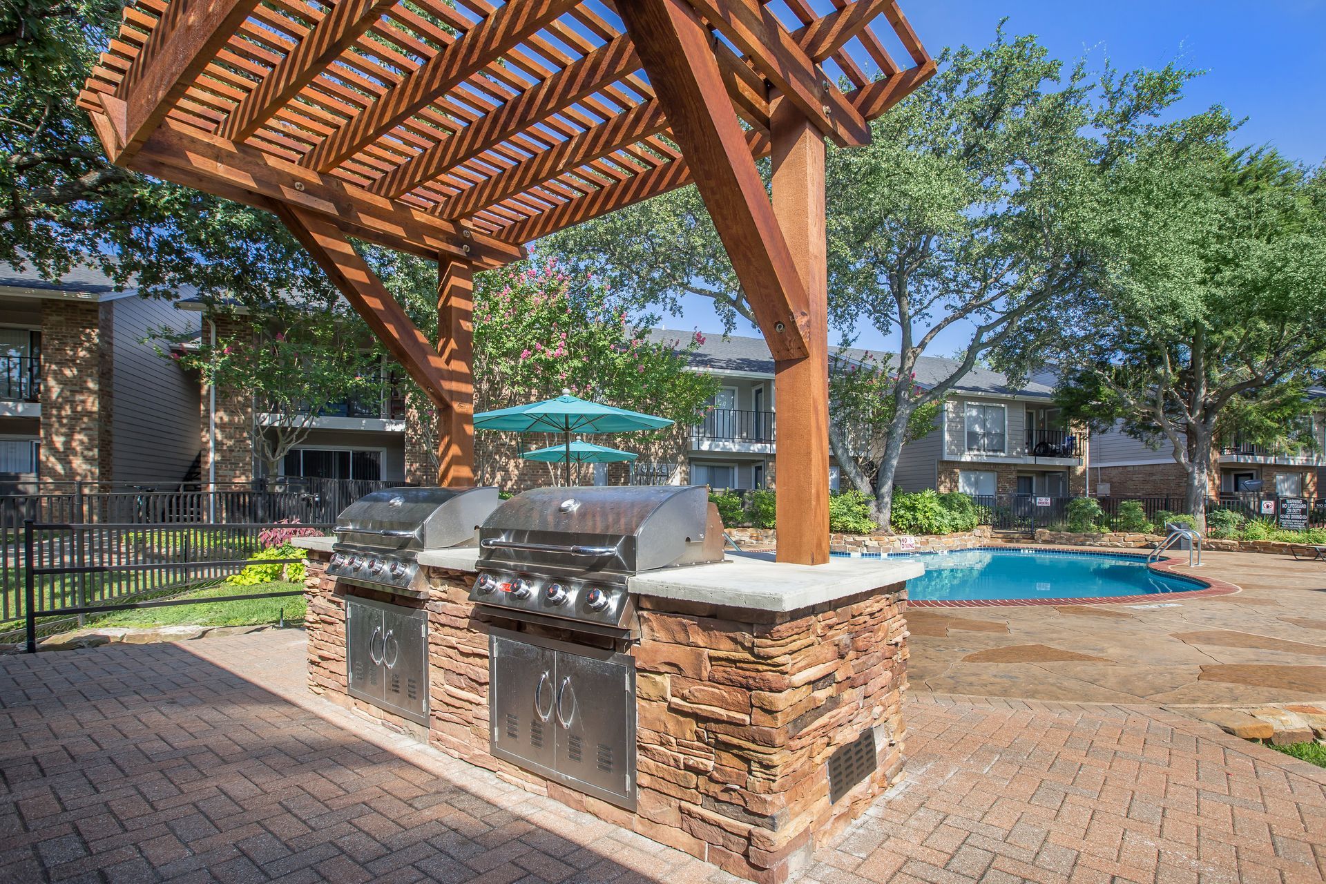 A grill is sitting under a wooden pergola next to a pool.