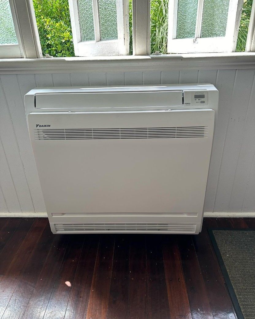 A White Air Conditioner is Sitting on a Wooden Floor Next to a Window — Eclipse Air Conditioning and Refrigeration In Redlynch, QLD