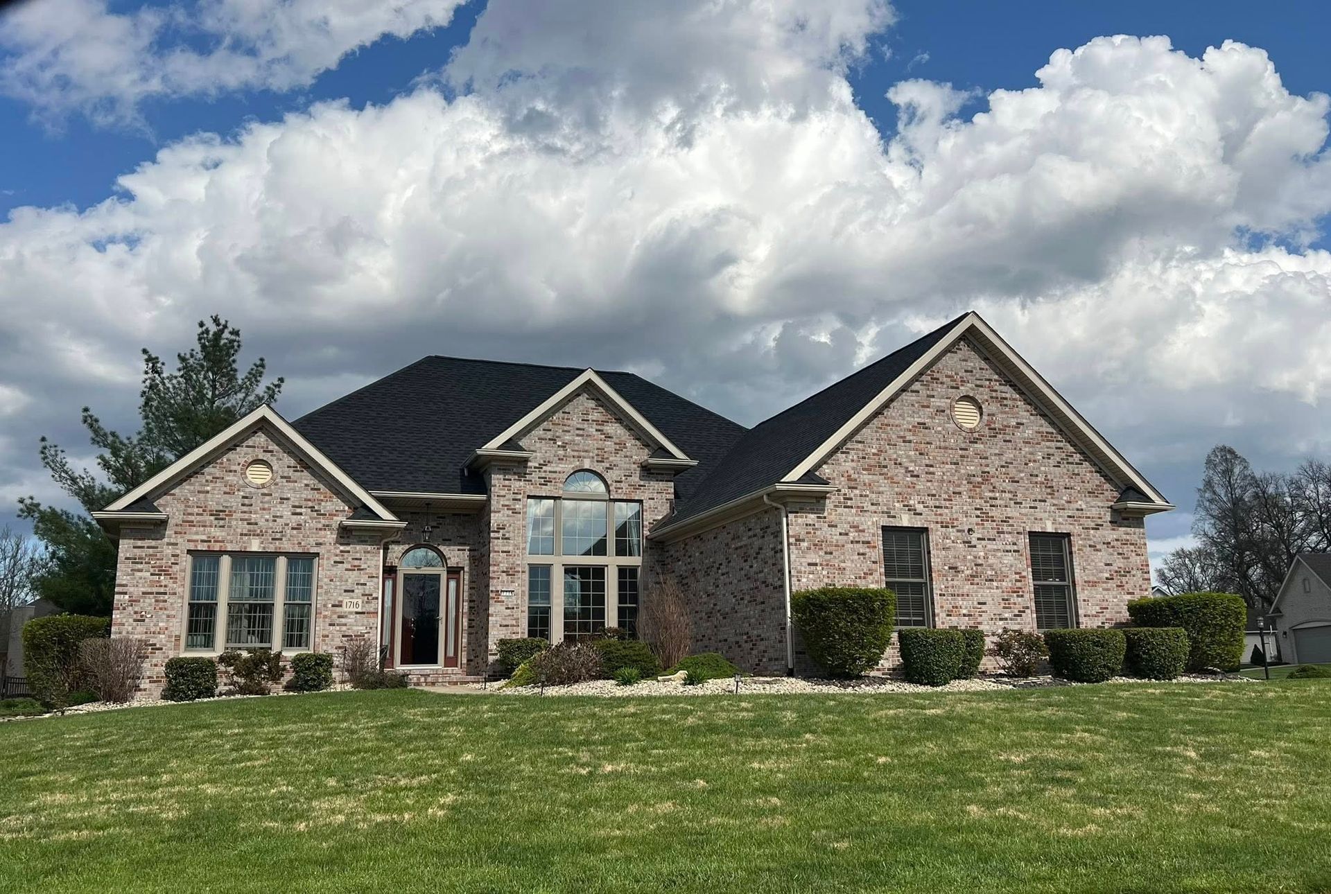 Brick house with dark roof and large front windows under a cloudy blue sky.