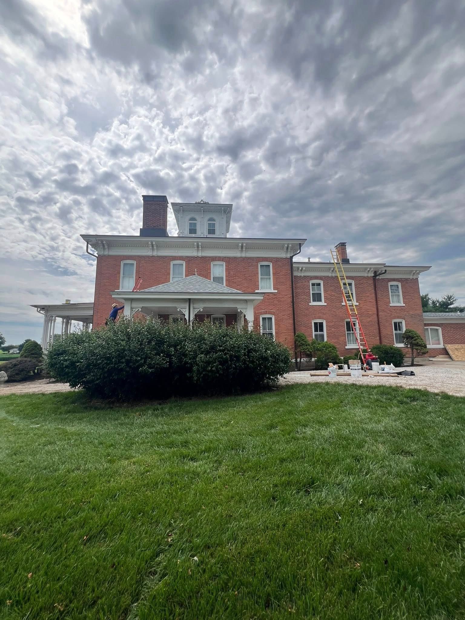 Red brick house with white trim under a cloudy sky. Workers are visible on the roof and a ladder is leaning against the building.