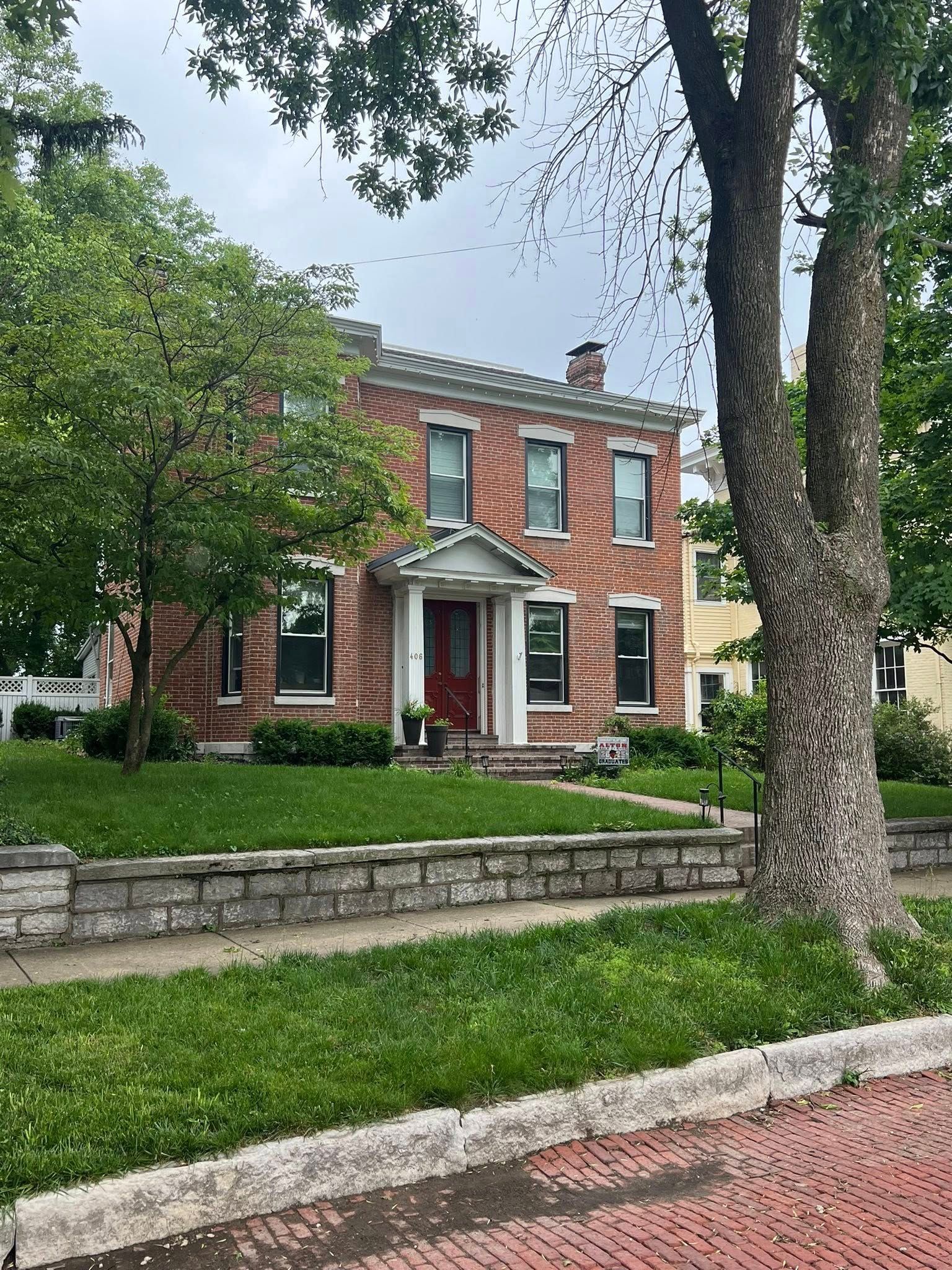 Red brick two-story house with white trim, green lawn, and stone wall. Tree in the foreground, cloudy sky.