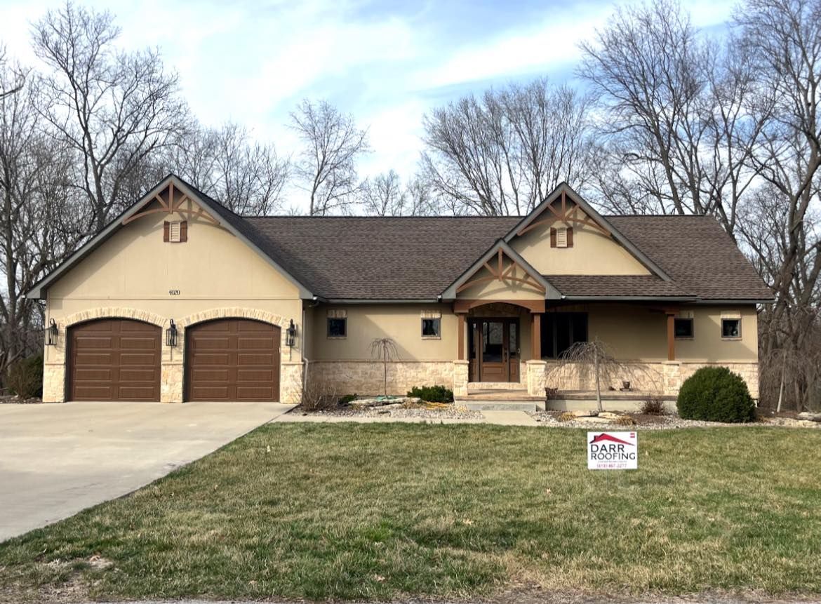 Tan stucco house with brown roof and garage doors, set in a grassy yard, with bare trees in the background.