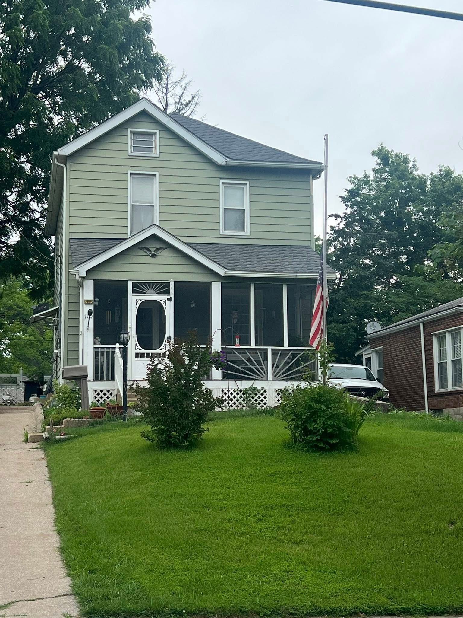 Two-story house with green siding, screened porch, American flag, and lawn.