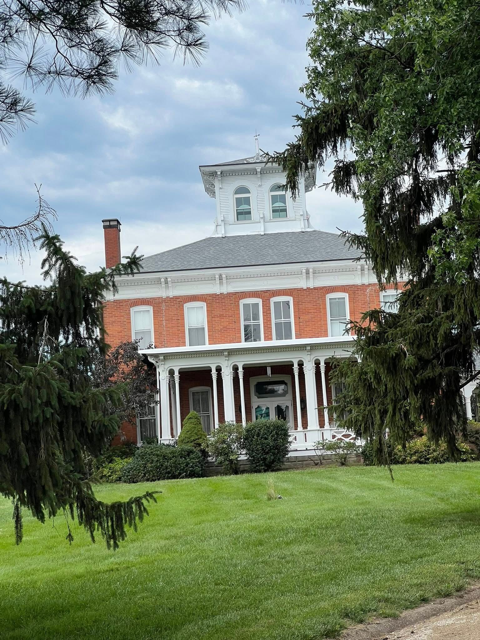 Red brick house with white trim, porch, and cupola on a green lawn, framed by trees under a cloudy sky.