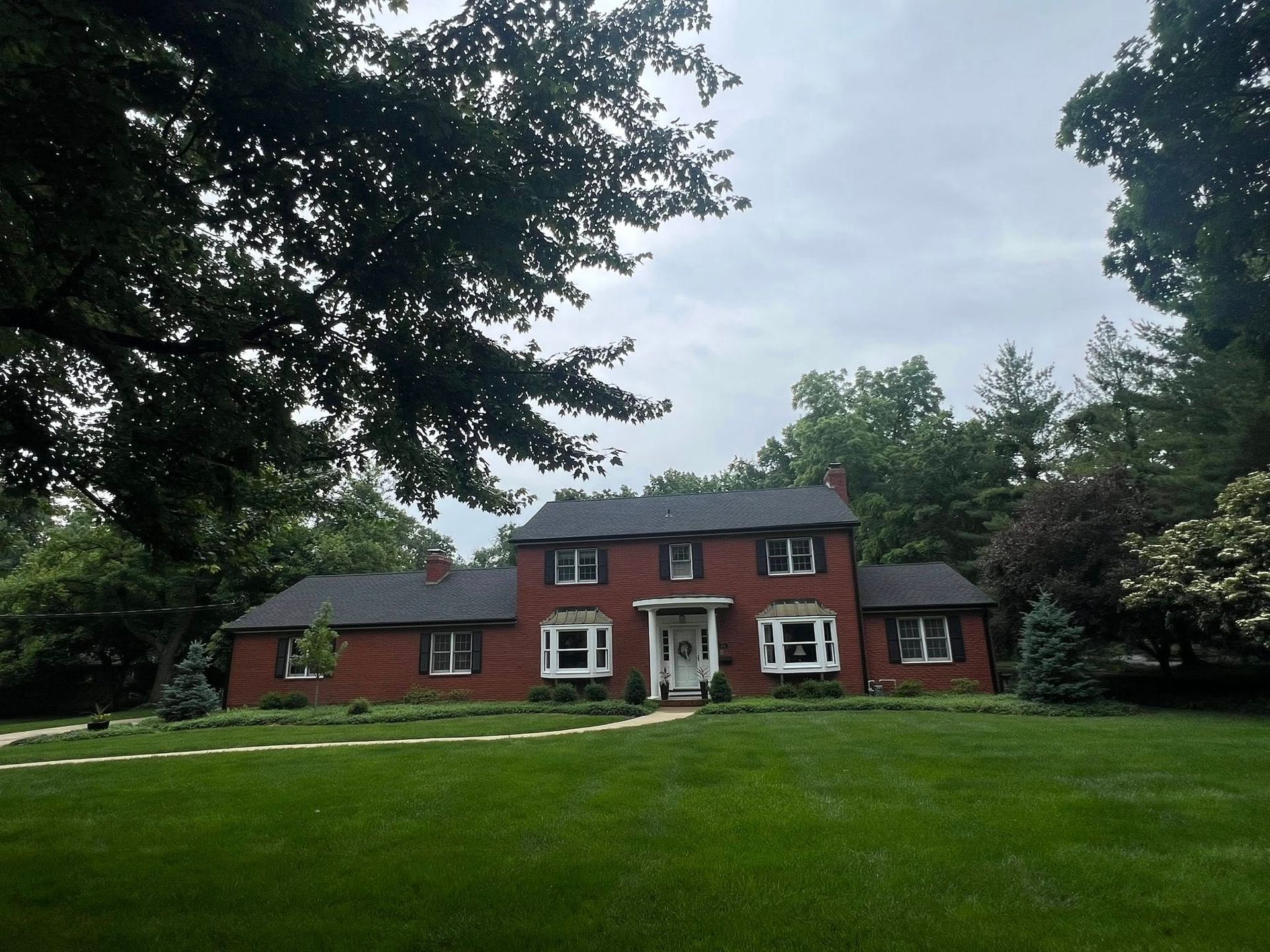 Red brick house with black roof, multiple windows, and a green lawn. Trees surround the house.