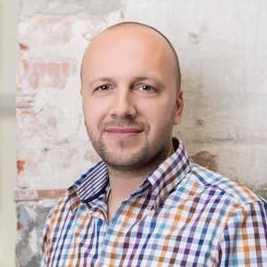 A person with a shaved head and light beard wearing a blue, white, and orange plaid shirt, posing against a brick wall.
