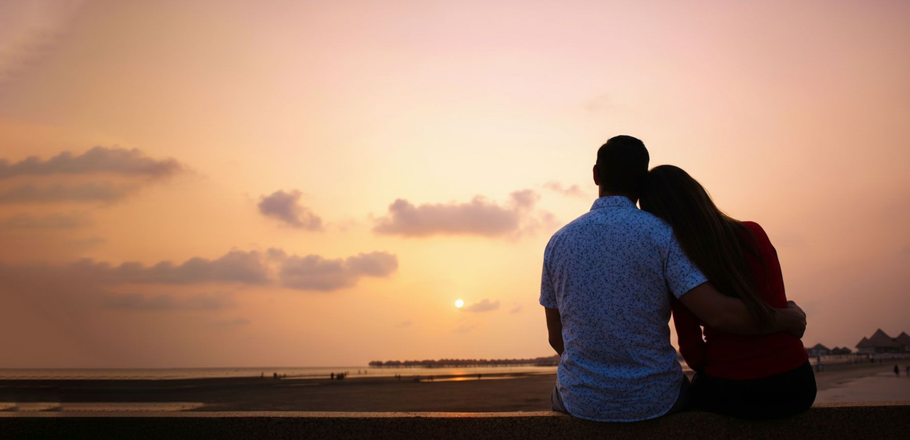 Couple sitting together on a beach at sunset, silhouetted against a warm orange sky.