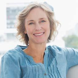 A person with shoulder-length blonde hair smiles while wearing a light blue textured top against a bright, blurred backdrop.