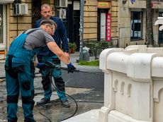 Two workers cleaning a white fountain with a power washer in a city setting.