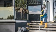 Man pressure washing outdoor steps, wearing yellow boots and gloves, next to a modern building.