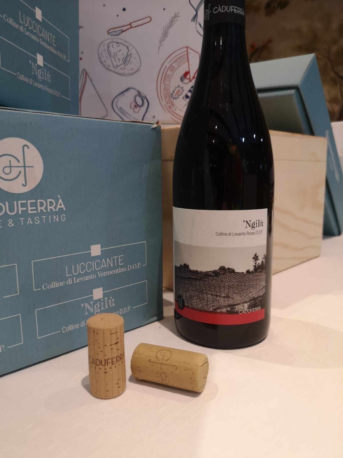 Un bicchiere di vino e una bottiglia su un tavolo. Il bicchiere è pieno di vino rosso. L'etichetta della bottiglia raffigura dei fiori blu.