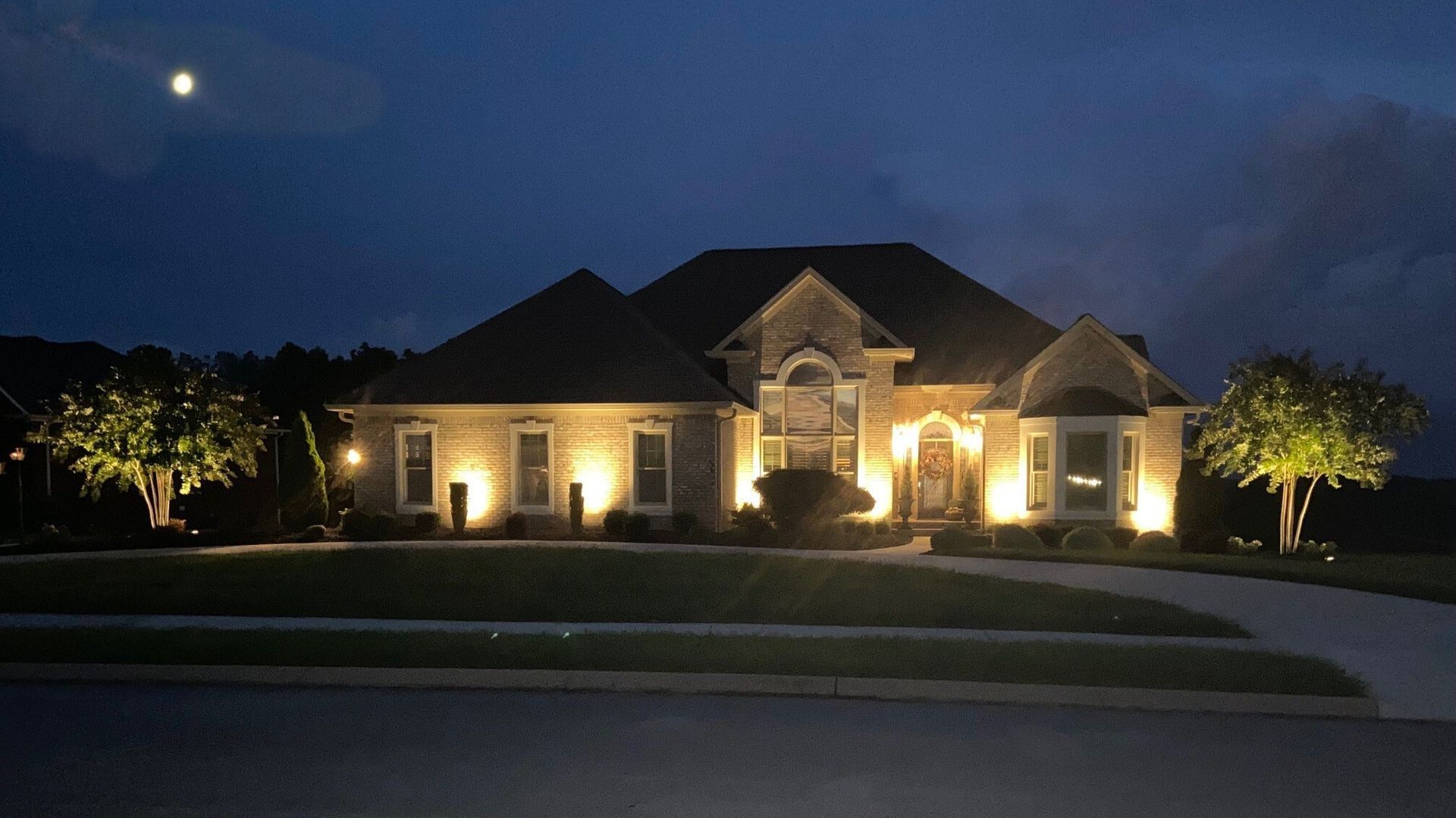 Single-story brick house warmly lit at night, with trees and a curved driveway in front.