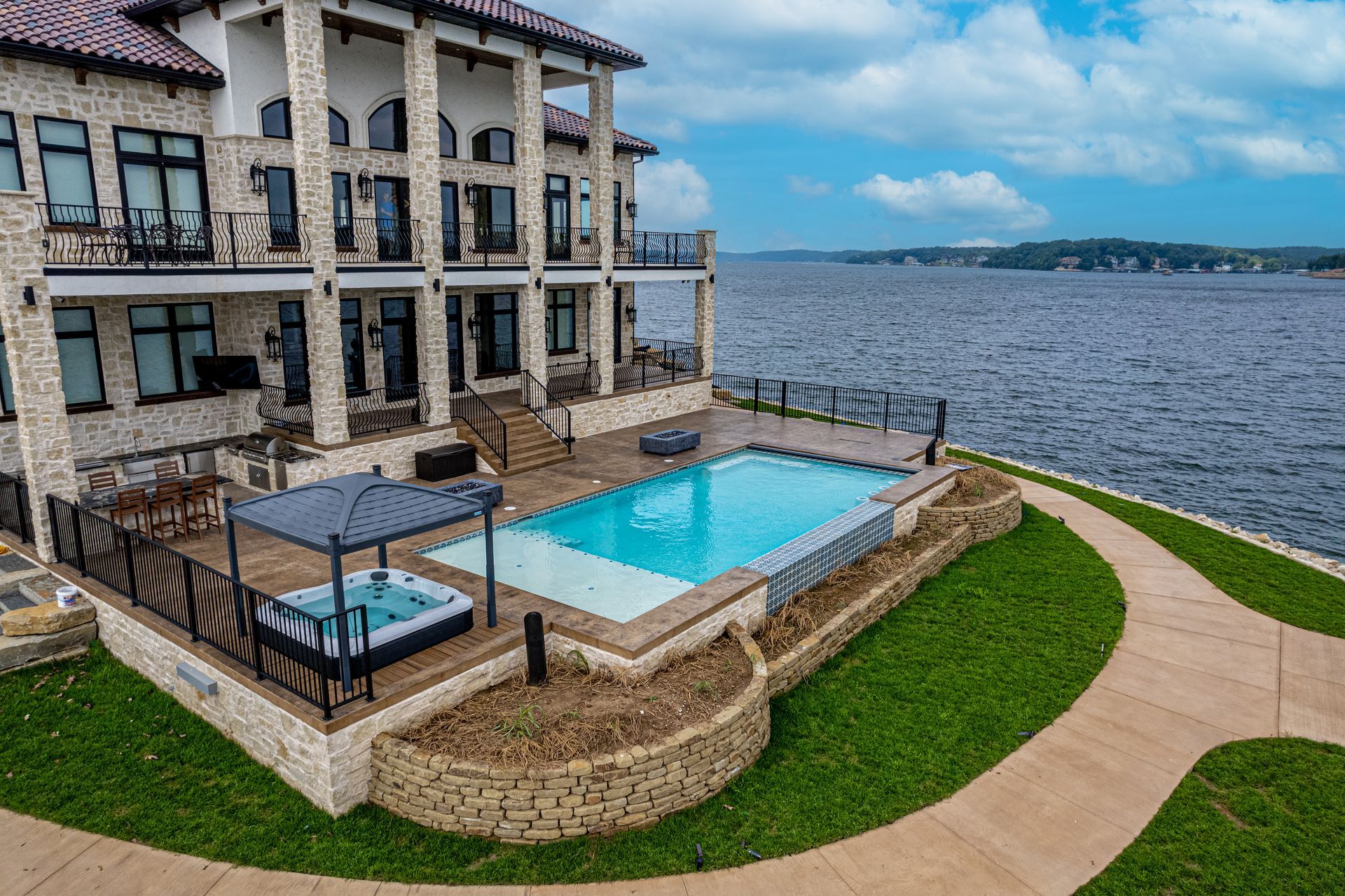 Pool overlooking a lake with a waterfall feature, two chairs on deck, and potted plants.