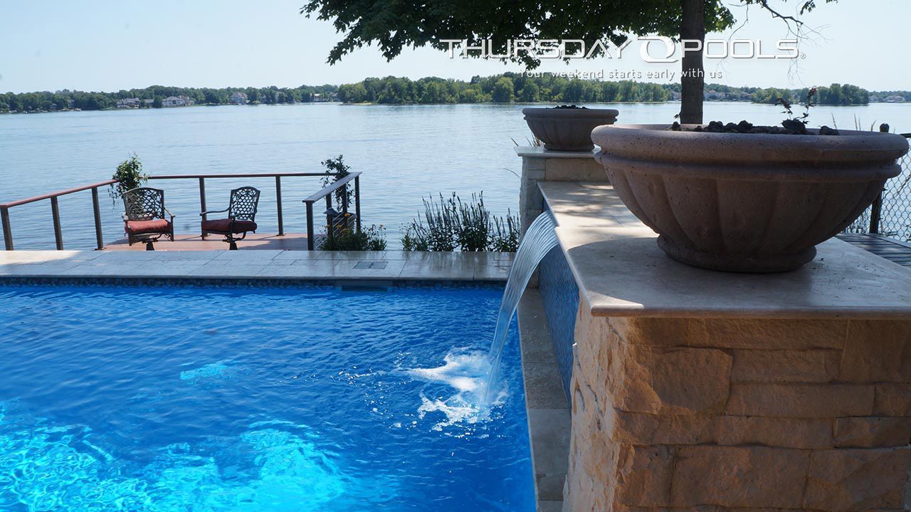 Pool overlooking a lake with a waterfall feature, two chairs on deck, and potted plants.